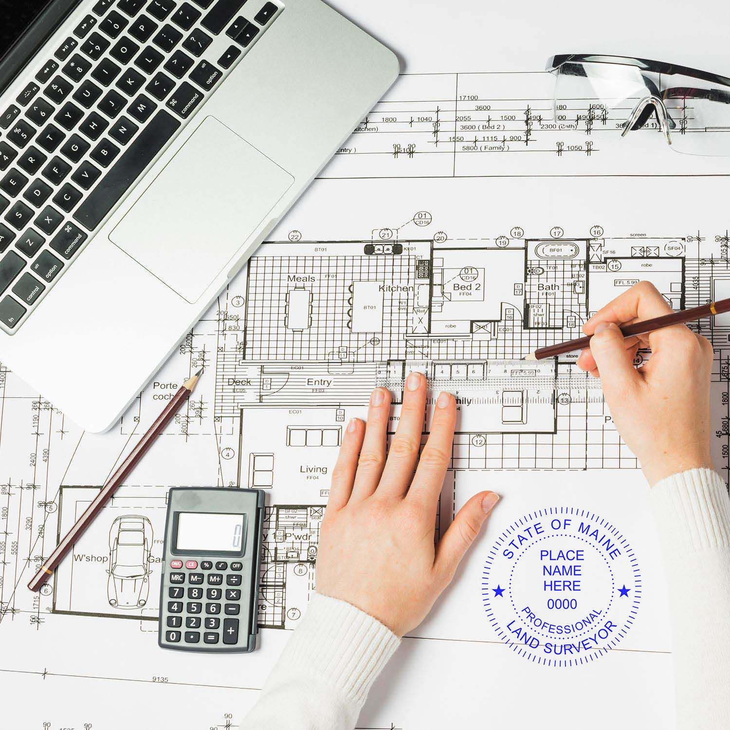 Person using Maine Land Surveyor Seal Stamp on architectural plans with a laptop, calculator, and glasses on the desk.