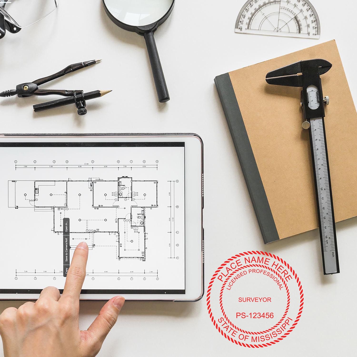 A desk with a tablet displaying a blueprint, compass, protractor, caliper, and a Self Inking Mississippi Land Surveyor Stamp on a notebook.