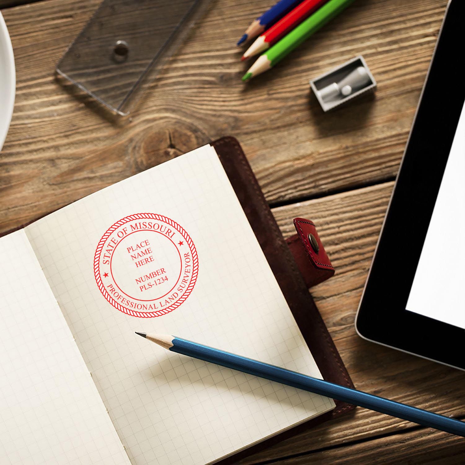 Missouri Land Surveyor Seal Stamp on an open notebook, surrounded by pencils, a sharpener, and a tablet on a wooden desk.