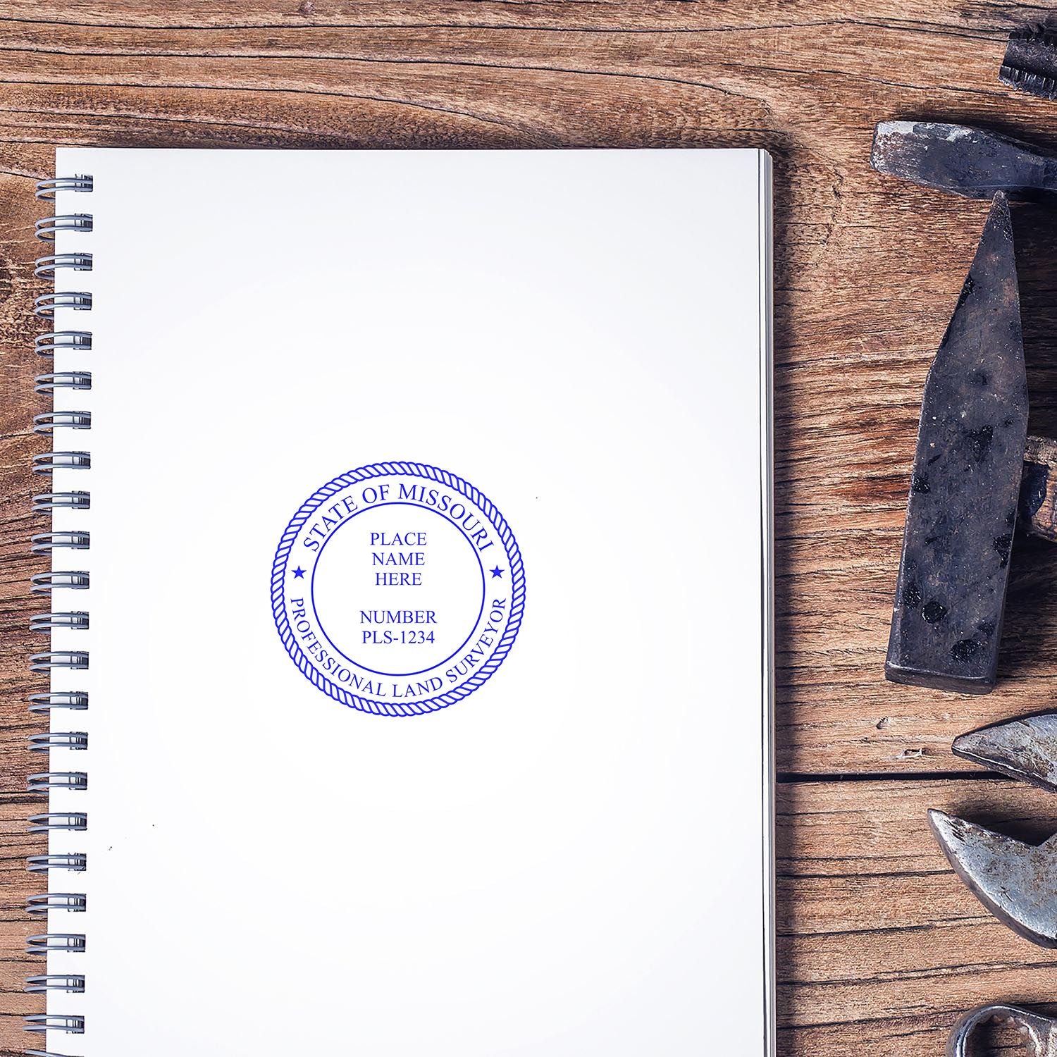 Self Inking Missouri Land Surveyor Stamp on a spiral notebook, surrounded by tools on a wooden surface.