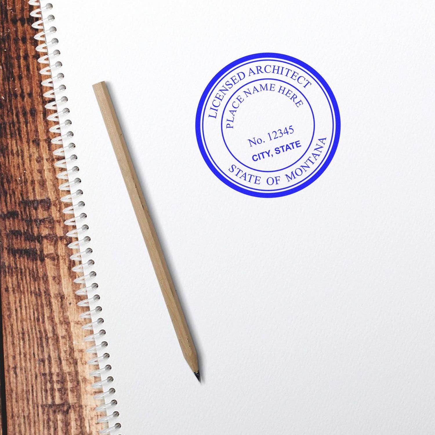 Self Inking Montana Architect Stamp imprint on white paper next to a pencil and spiral notebook on a wooden surface.