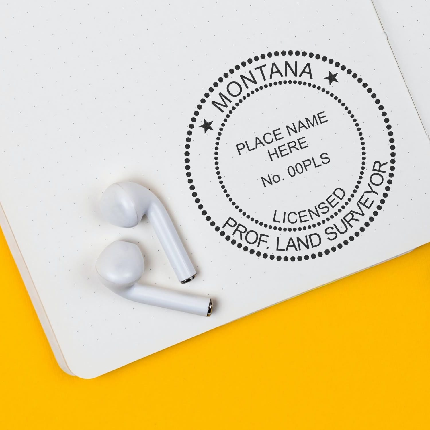 Premium MaxLight Pre-Inked Montana Surveyors Stamp on a notebook with two white earbuds on a yellow background.