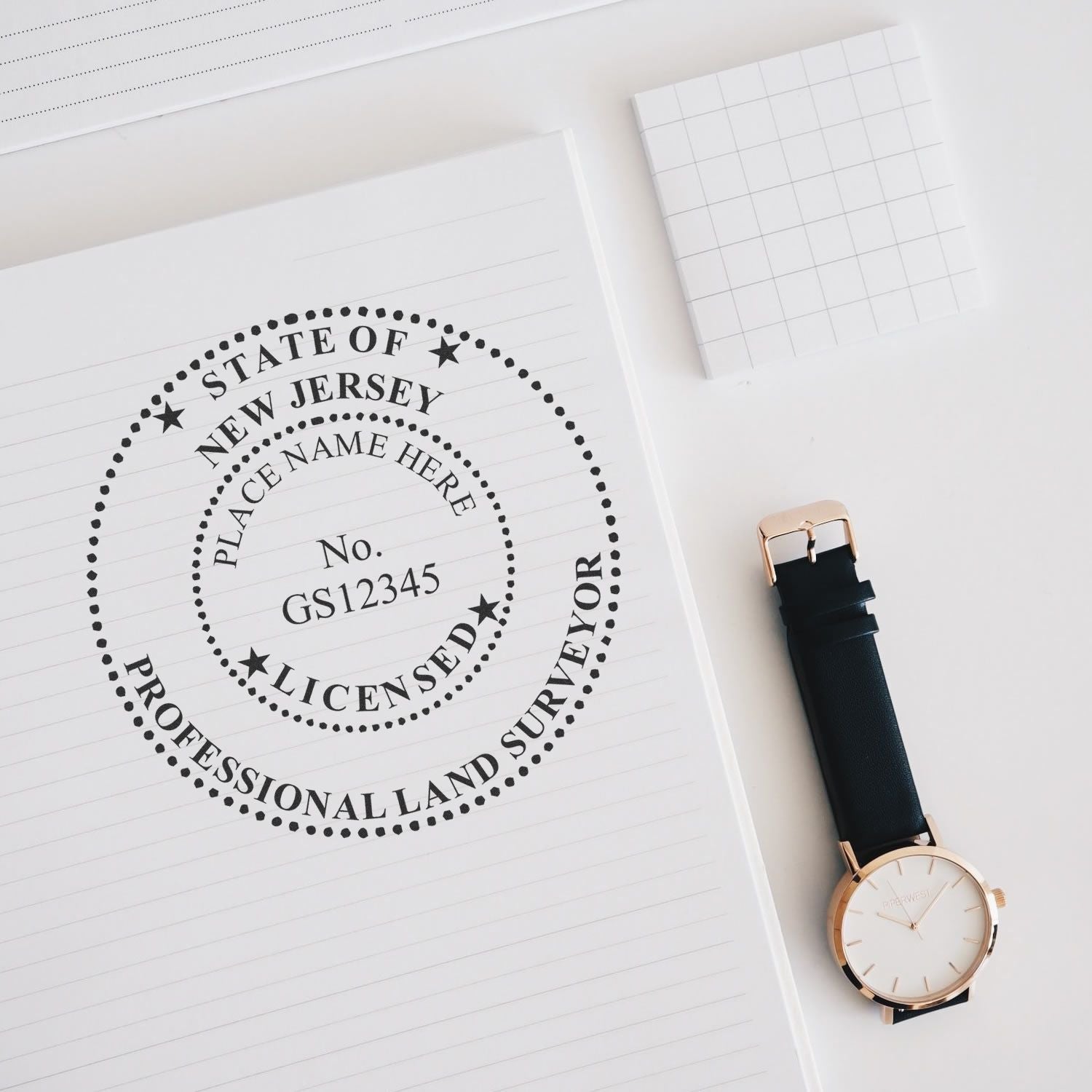 Self Inking New Jersey Land Surveyor Stamp on a notepad with a watch and a small grid notebook nearby.