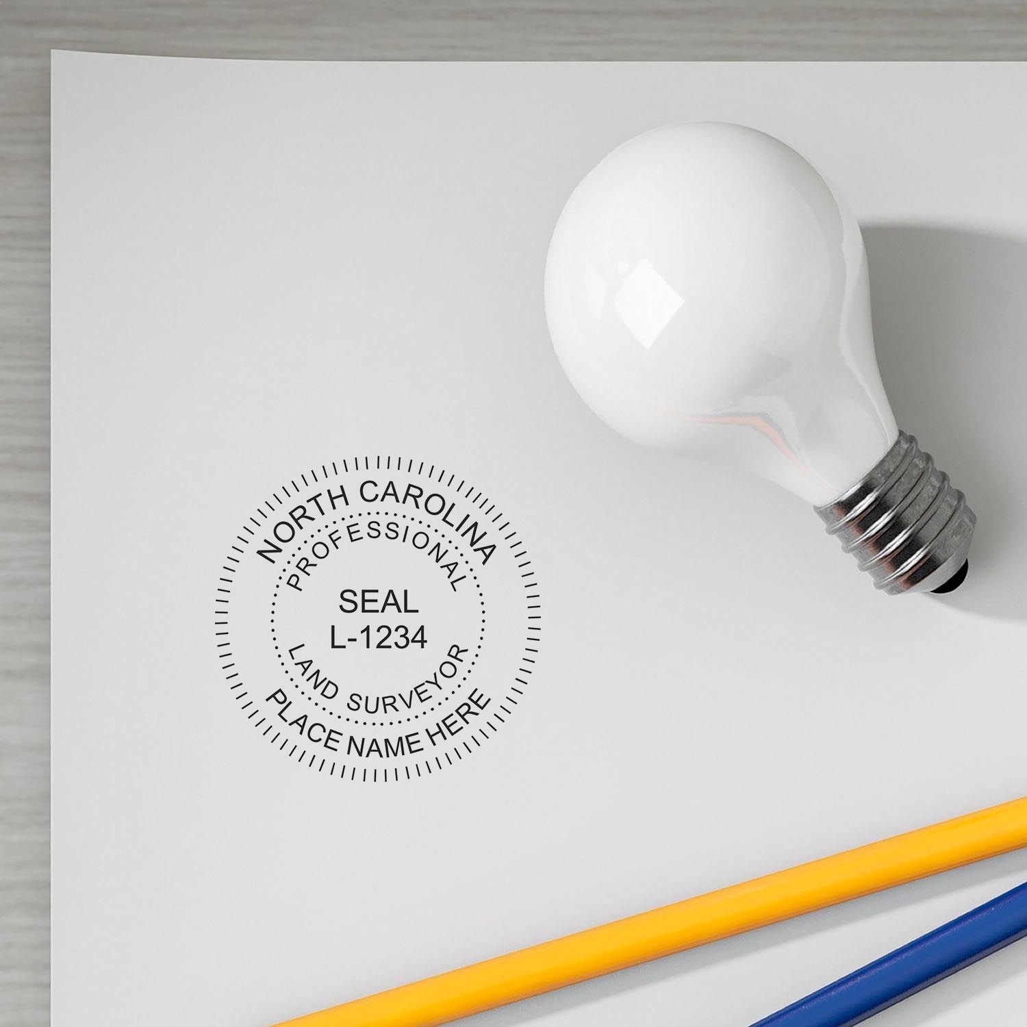 Self Inking North Carolina Land Surveyor Stamp on white paper with a light bulb and pencils nearby.