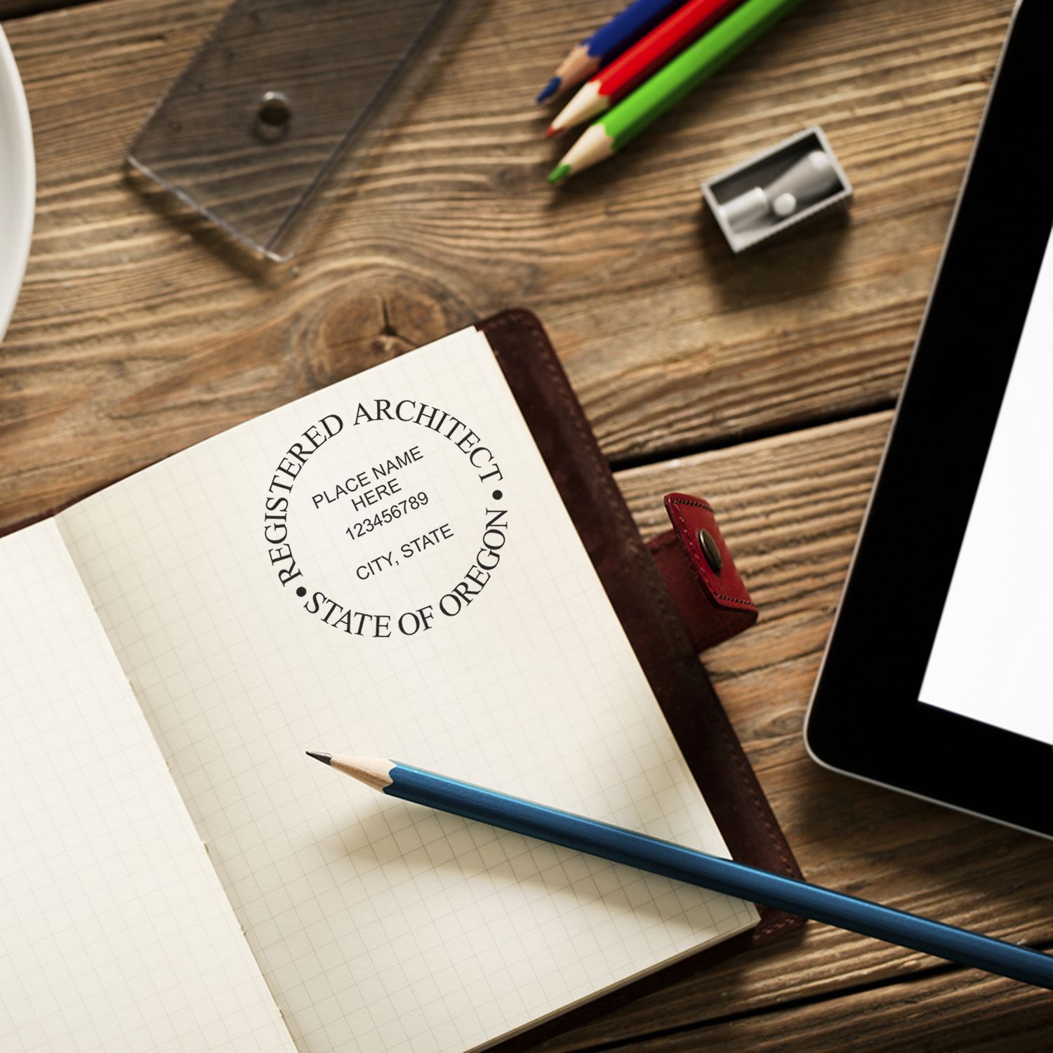 Self Inking Oregon Architect Stamp used on a notebook, surrounded by pencils, a sharpener, and a tablet on a wooden desk.