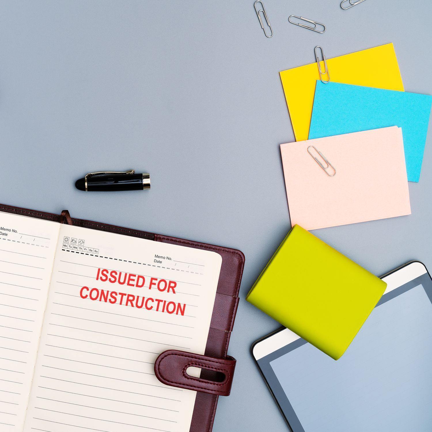 Desk with a planner stamped 'ISSUED FOR CONSTRUCTION' using a Regular Issued for Construction Stamp, surrounded by office supplies.