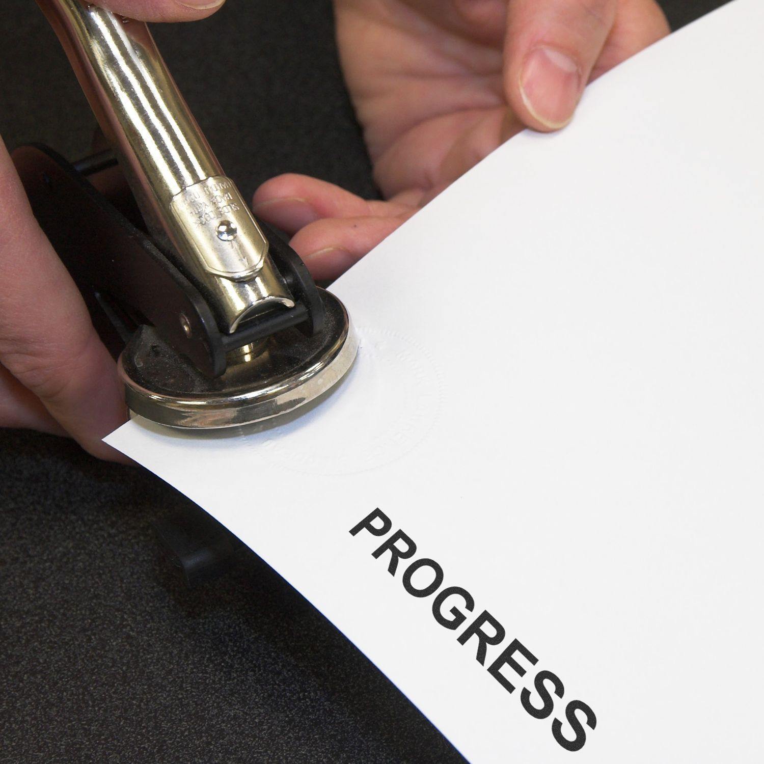 Person using a Slim Pre-Inked Progress Stamp on a white sheet of paper with the word PROGRESS stamped in bold black letters.