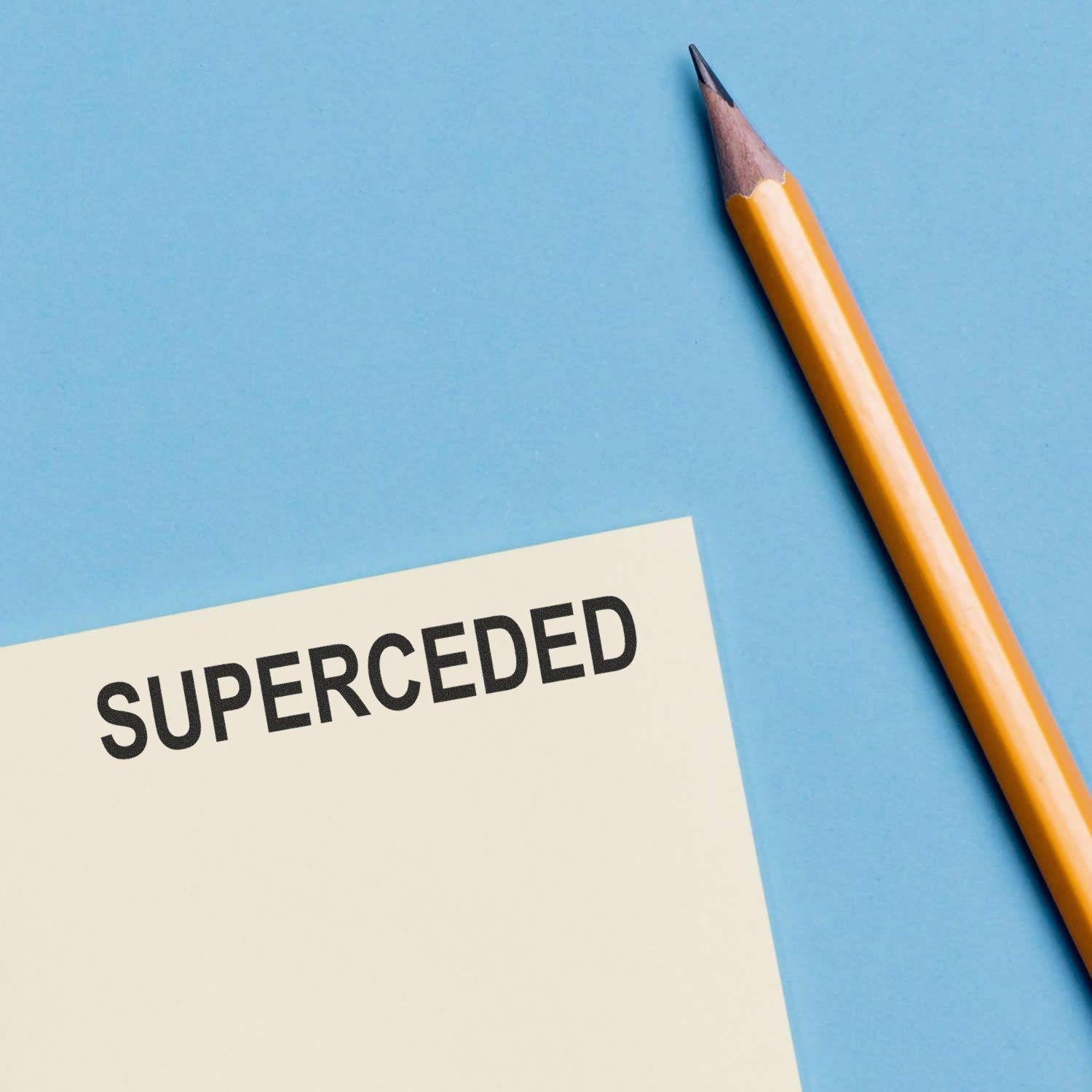 Slim Pre-Inked Superceded Stamp marking SUPERCEDED on paper next to a yellow pencil on a blue background.