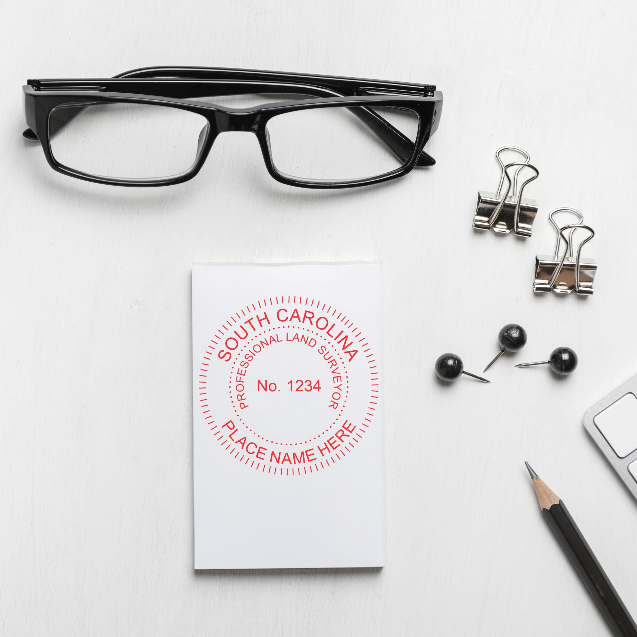 South Carolina Land Surveyor Seal Stamp, SC PLS Stamp on a white paper beside glasses, binder clips, push pins, pencil, and keyboard.