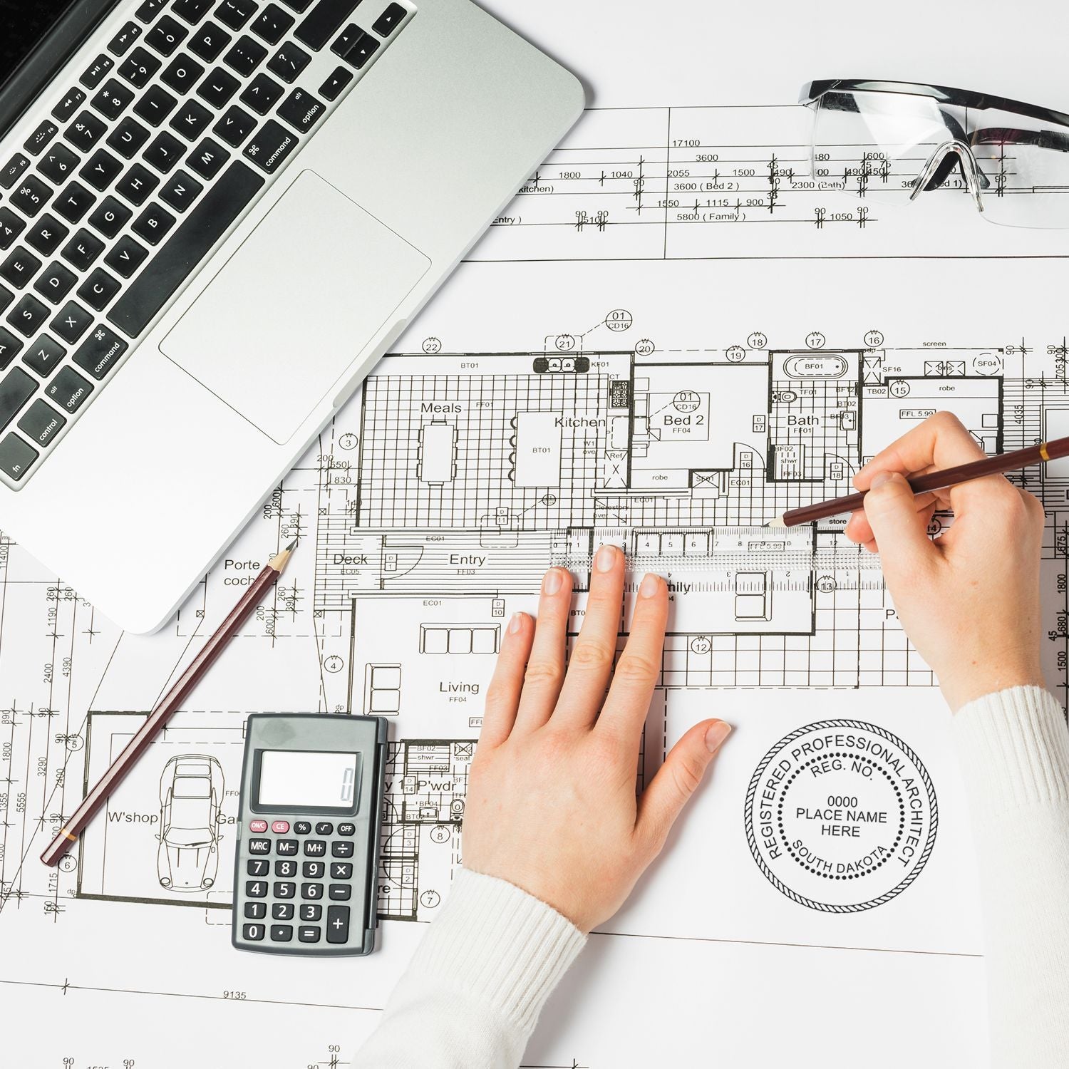 Person using Self Inking South Dakota Architect Stamp on architectural blueprints, with a laptop, calculator, and drafting tools nearby.