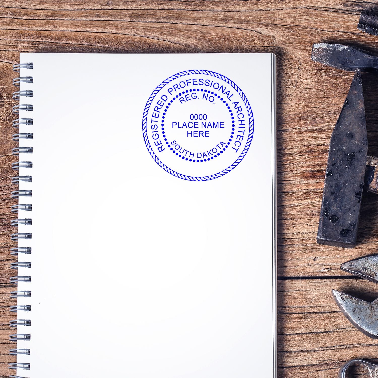 Self Inking South Dakota Architect Stamp in blue ink on a spiral notebook, placed on a wooden desk with tools nearby.