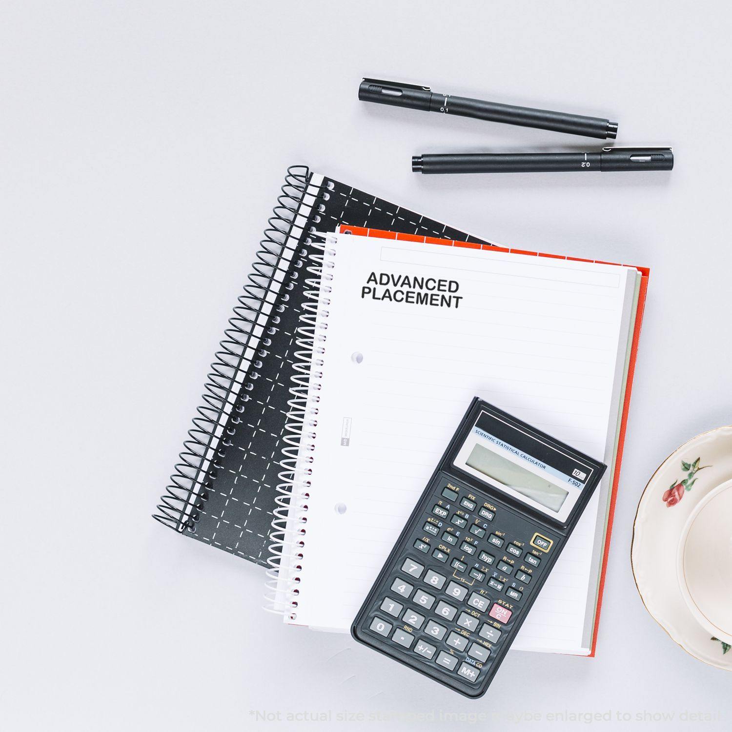 Large Advanced Placement Rubber Stamp used on a notebook, with a calculator, pens, and a plate on a white surface.