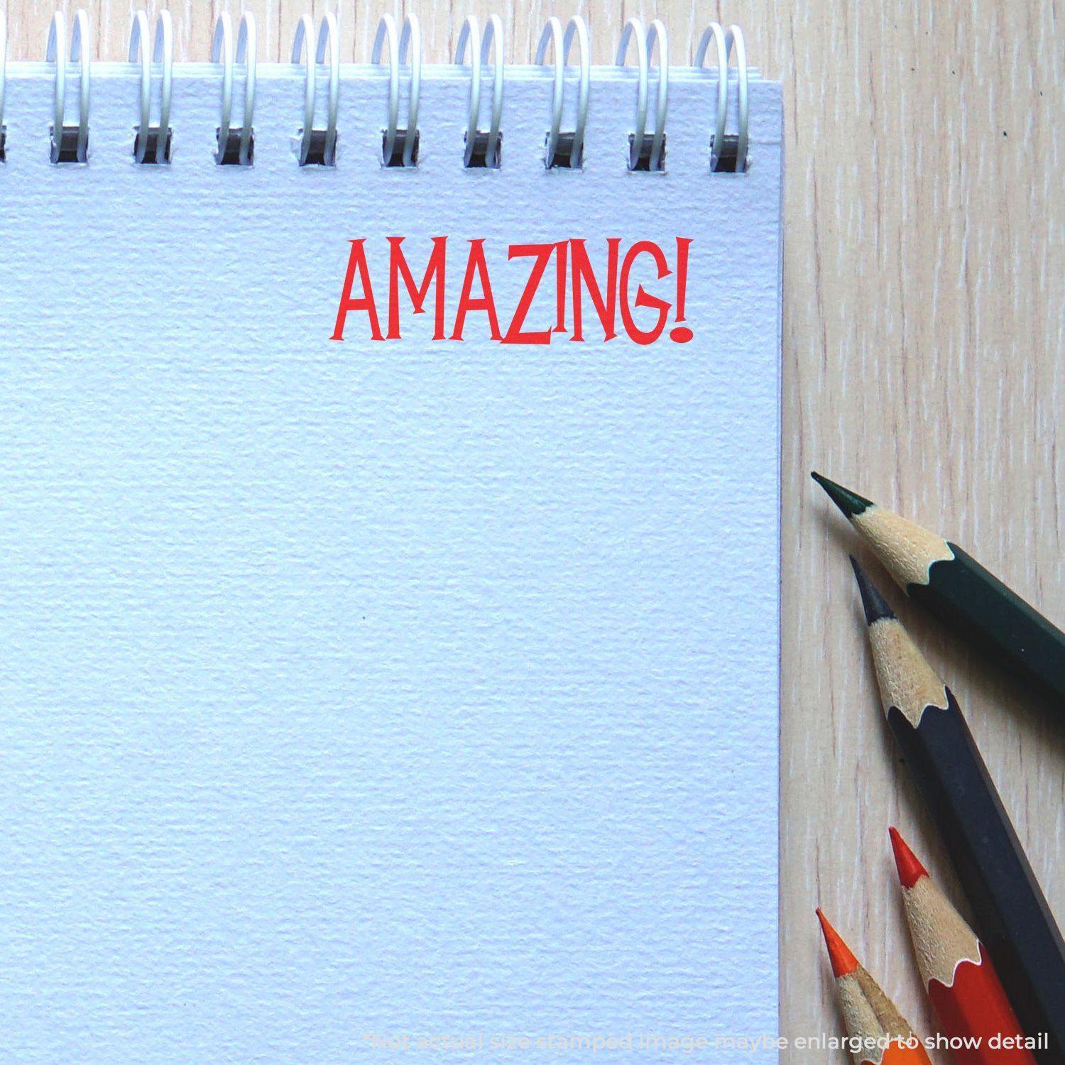 A Large Self Inking Amazing Stamp imprinting the word AMAZING! in red ink on a spiral-bound notebook, with colored pencils nearby.