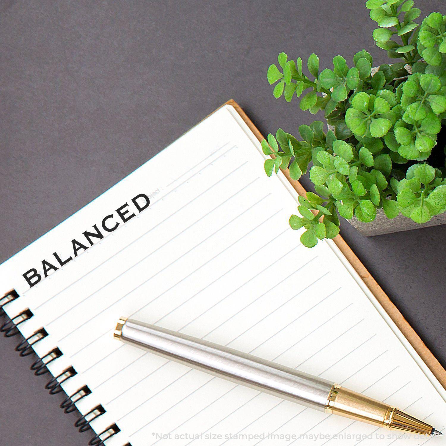 Balanced rubber stamp on a notebook page with a pen and a small potted plant on a gray surface.