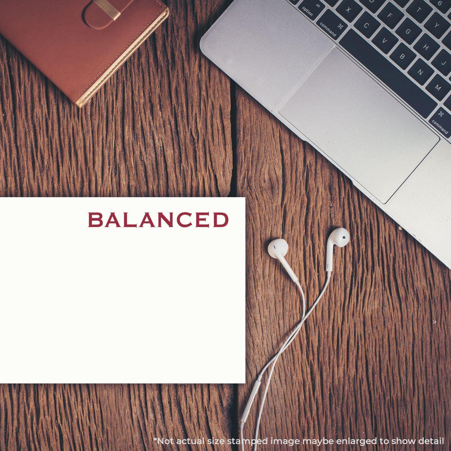 A Balanced rubber stamp on a white card, placed on a wooden desk with a laptop, notebook, and earphones nearby.