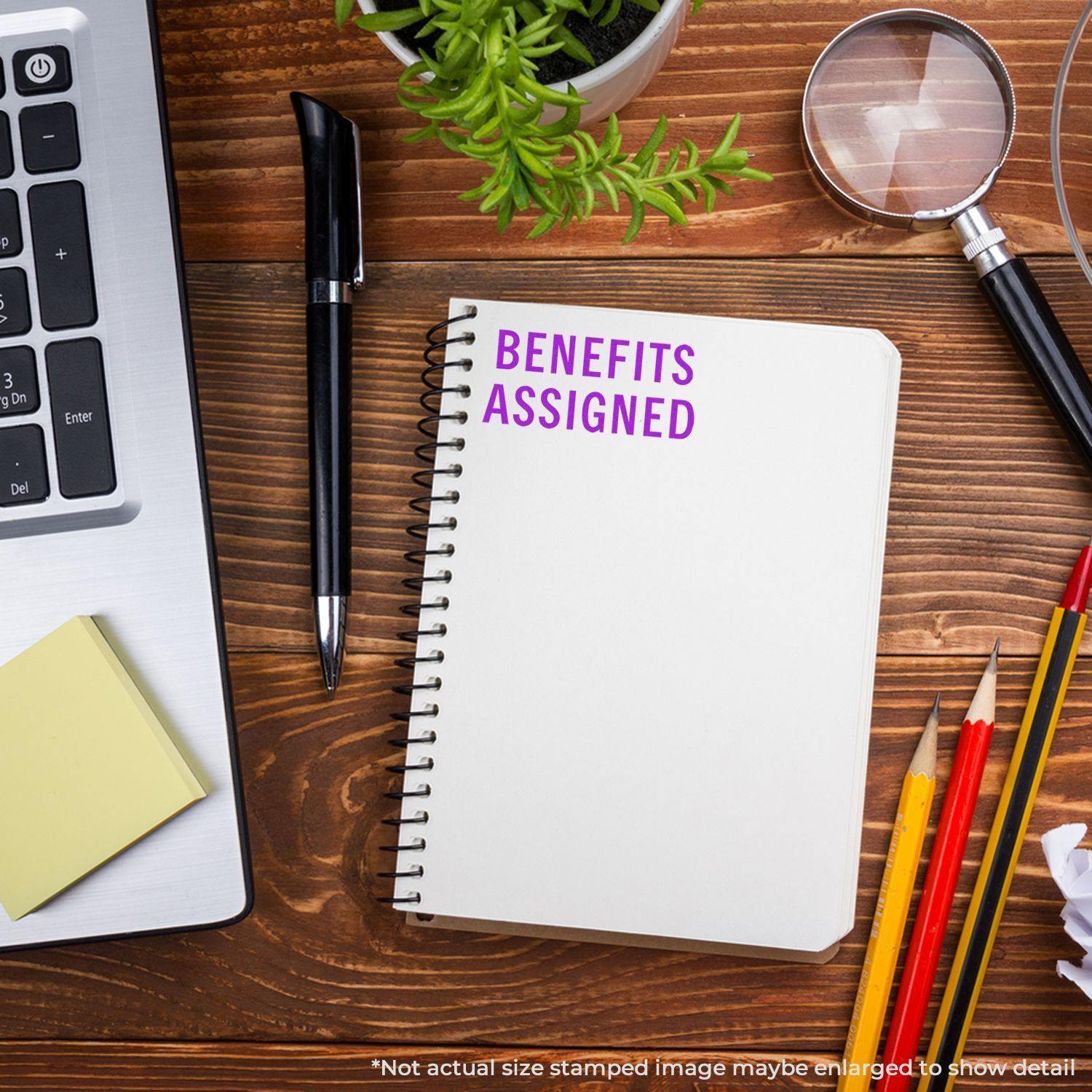 A Large Benefits Assigned Rubber Stamp is used on a spiral notebook placed on a wooden desk with office supplies and a plant.