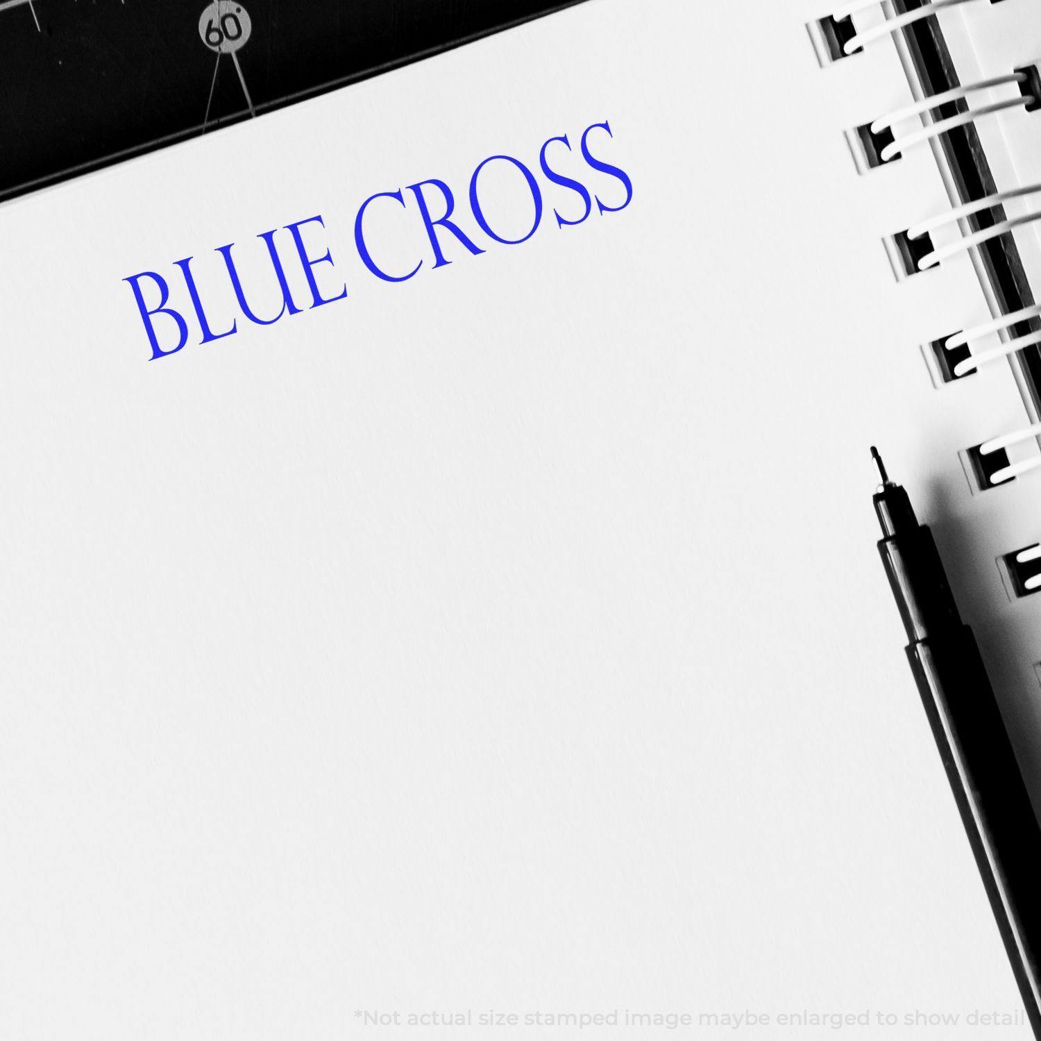 Large Pre-Inked Blue Cross Stamp in use on white paper, showing a clear blue BLUE CROSS imprint next to a black pen and spiral notebook.