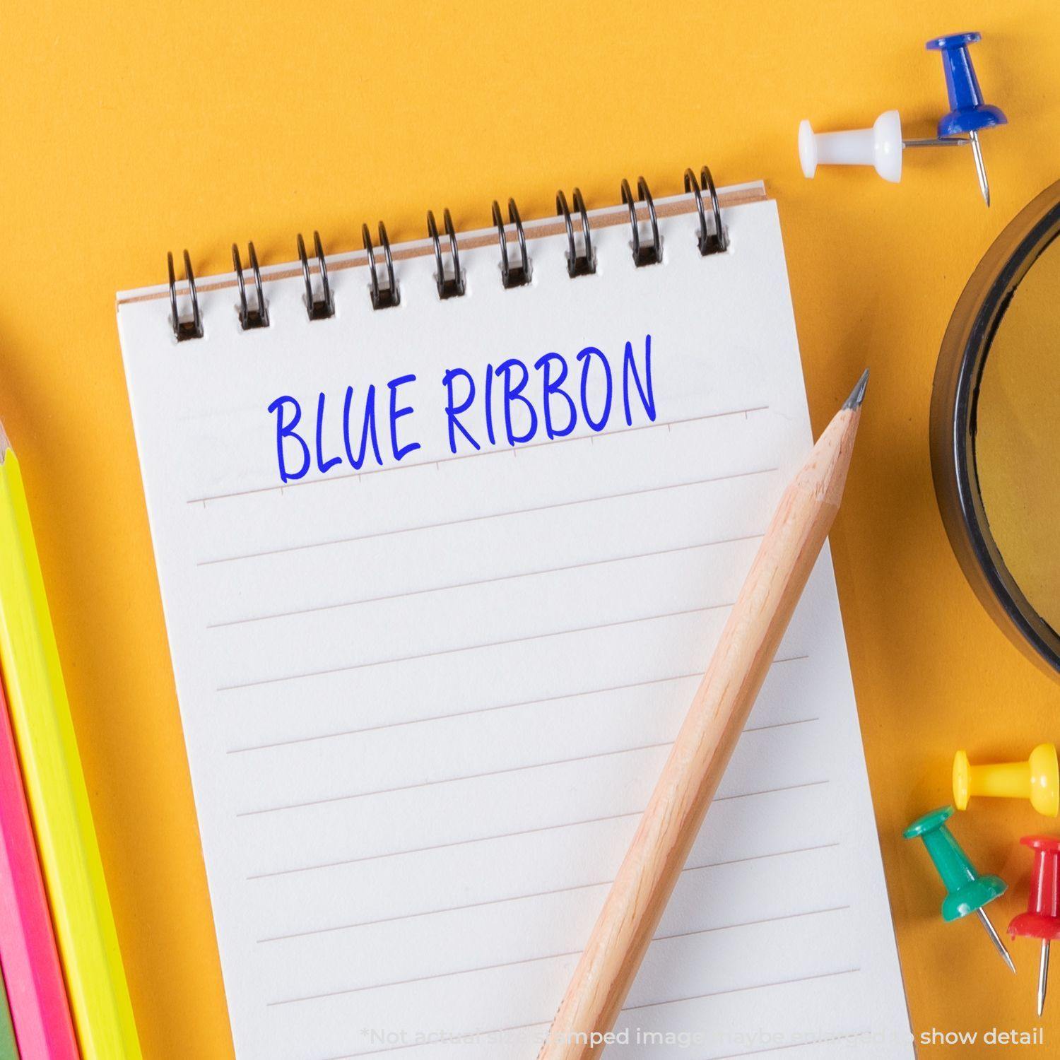 A Large Blue Ribbon Rubber Stamp is used on a notepad with a pencil, colorful push pins, and a yellow background.