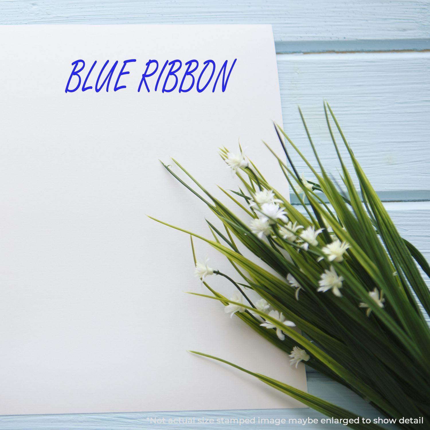 Blue Ribbon Rubber Stamp impression on white paper next to green grass and white flowers on a light blue wooden surface.