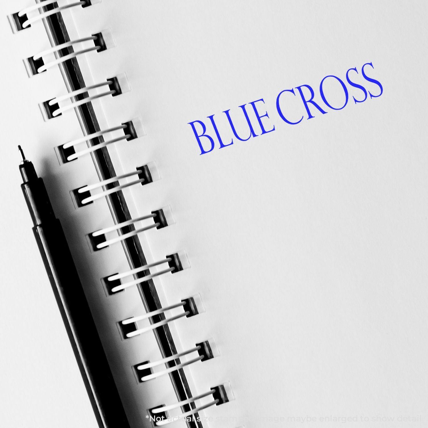 Large Pre-Inked Blue Shield Stamp imprinting BLUE CROSS on a white notebook page with a black pen beside it.