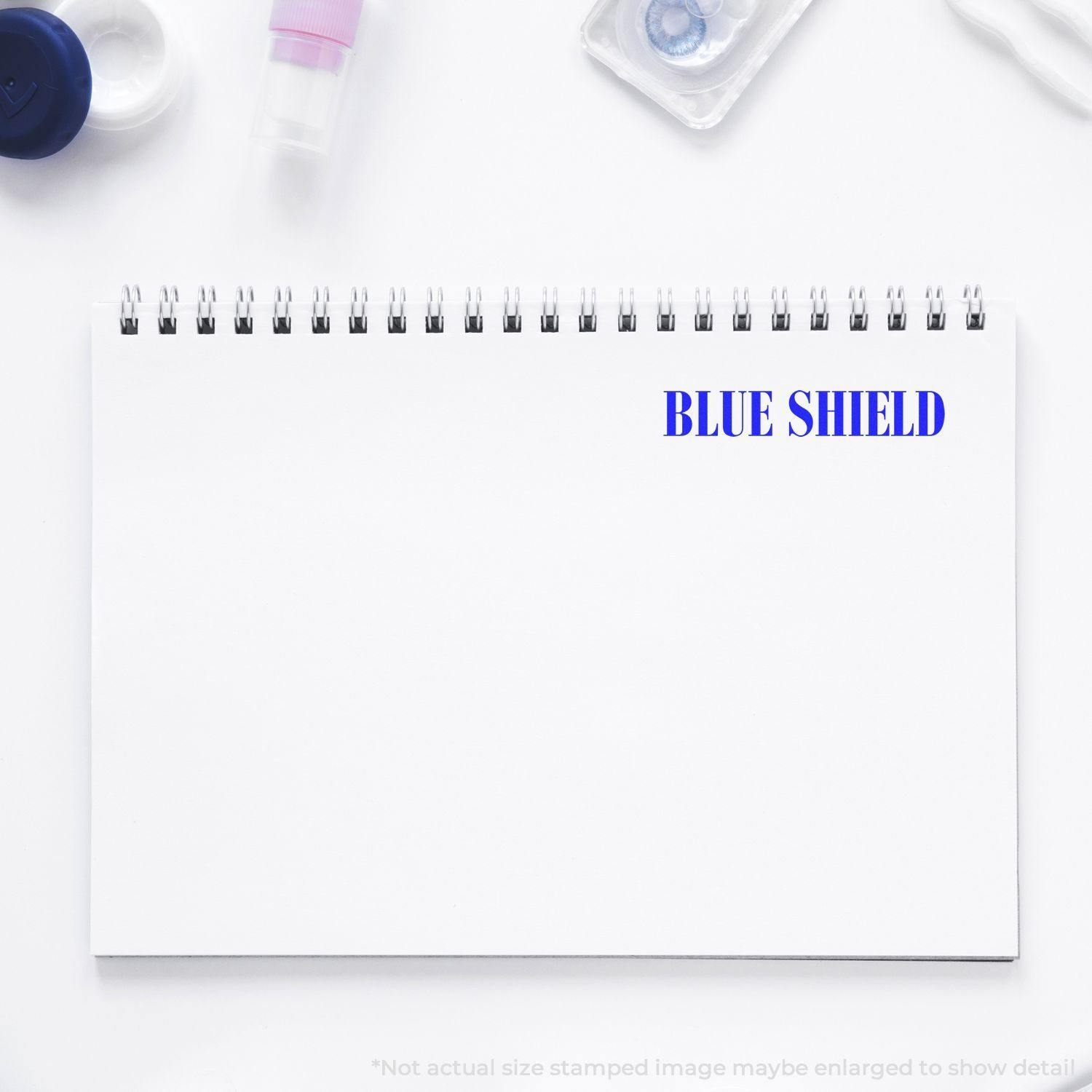 Large Blue Shield Rubber Stamp imprint on a white spiral notebook, surrounded by office supplies.