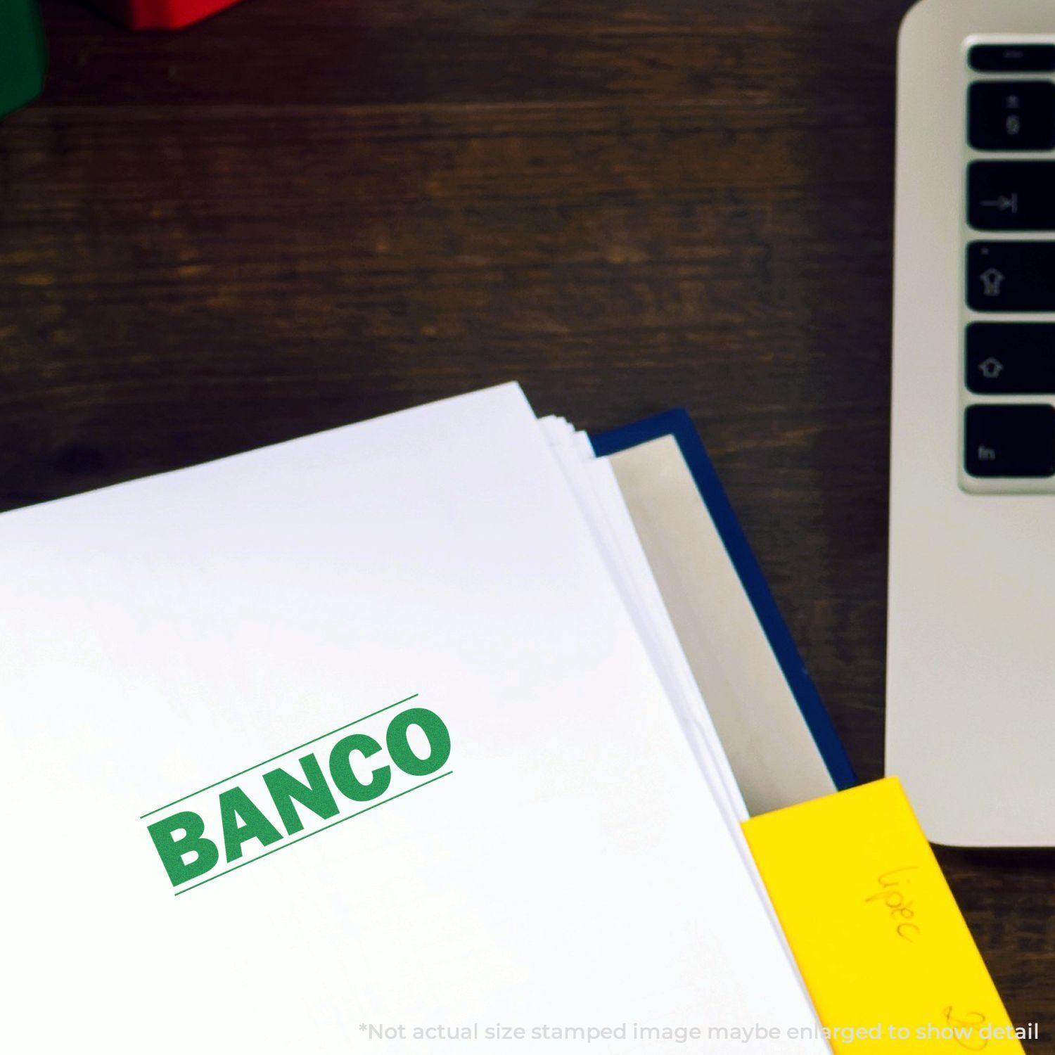 Large Pre-Inked Bold Banco Stamp imprint on a white document near a laptop and yellow sticky note on a wooden desk.