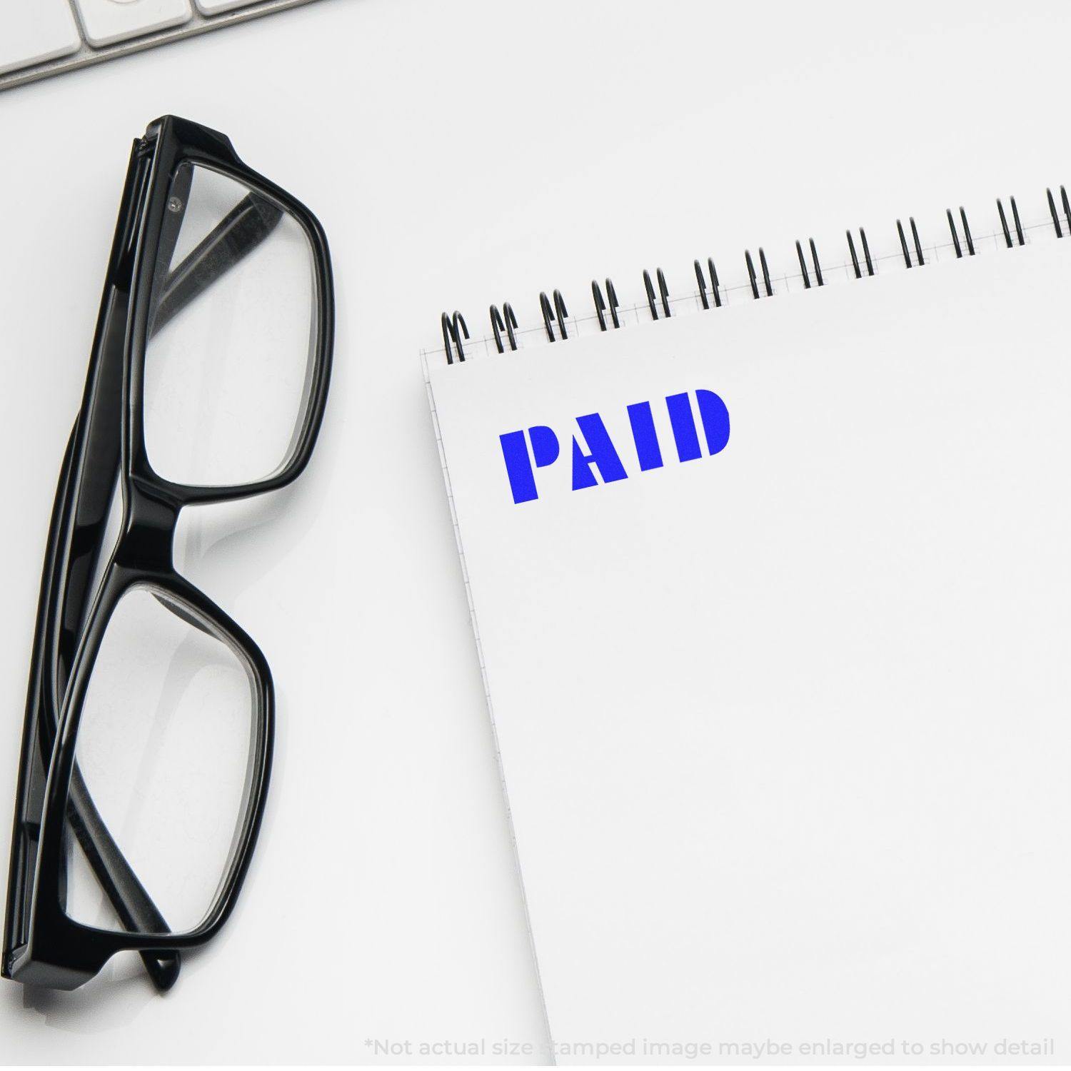 A pair of black glasses next to a notebook with a bold PAID rubber stamp in blue ink on a white page.