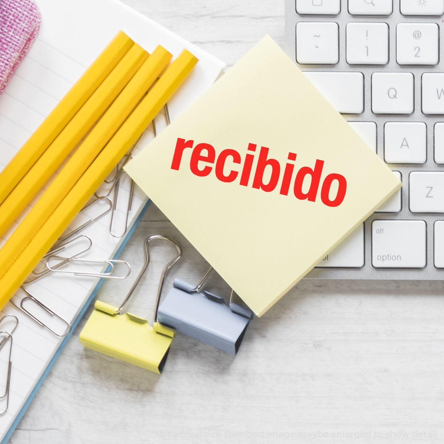 Self Inking Bold Recibido Stamp on a yellow sticky note, surrounded by pencils, paper clips, binder clips, and a keyboard.