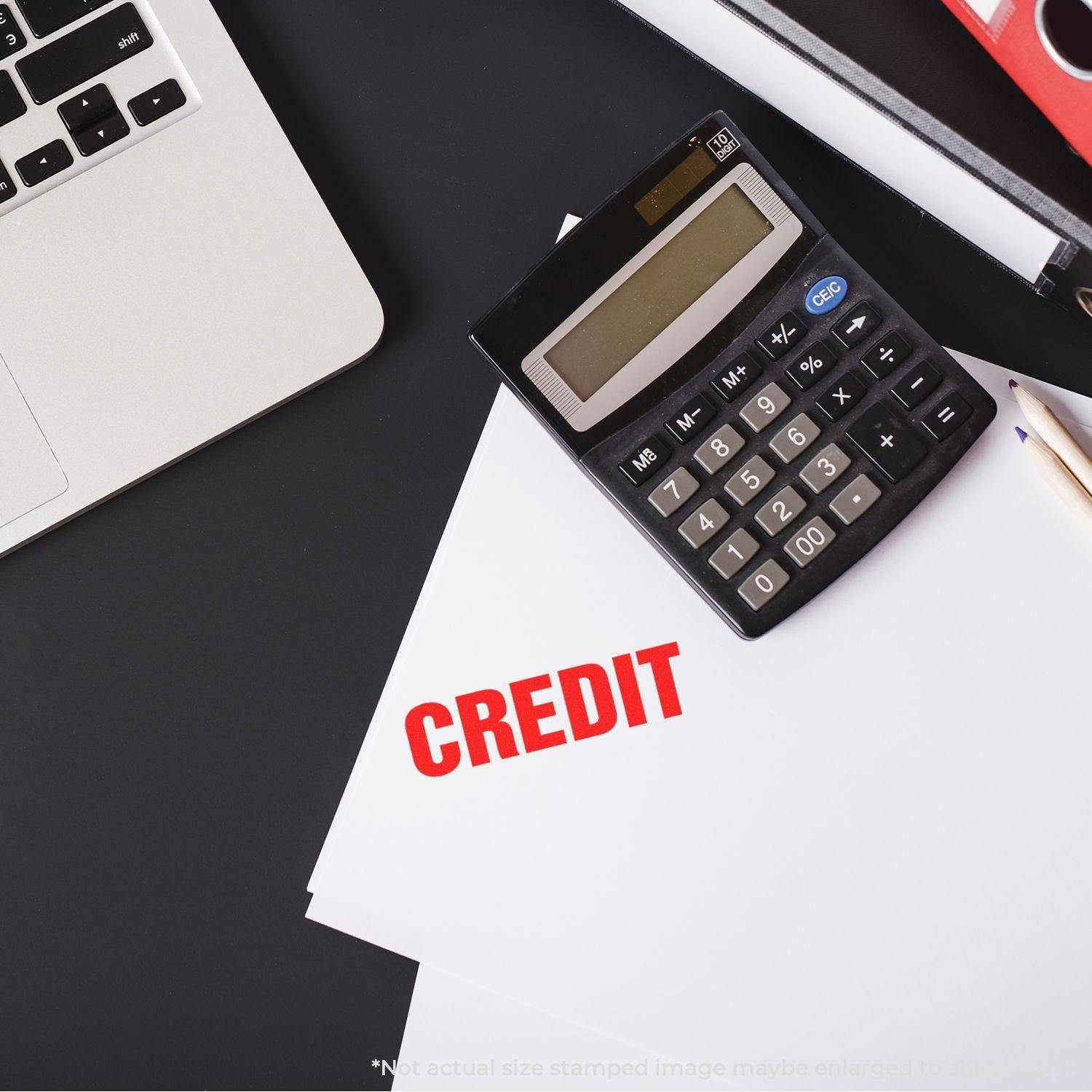 Self Inking Bold Red Credit Stamp marking CREDIT on paper, next to a calculator, pencil, and laptop on a black desk.