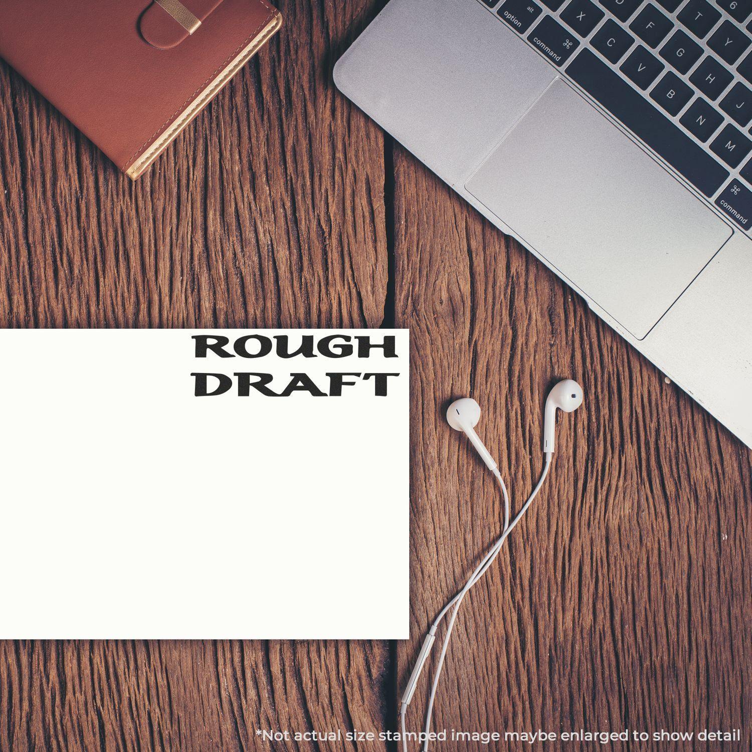 Large Pre-Inked Bold Rough Draft Stamp used on paper, placed on a wooden desk next to a laptop, notebook, and earphones.