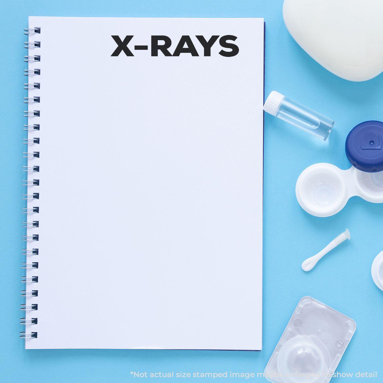 Bold X-Rays rubber stamp on a white notebook page, surrounded by contact lens case, solution bottle, and cotton swab.