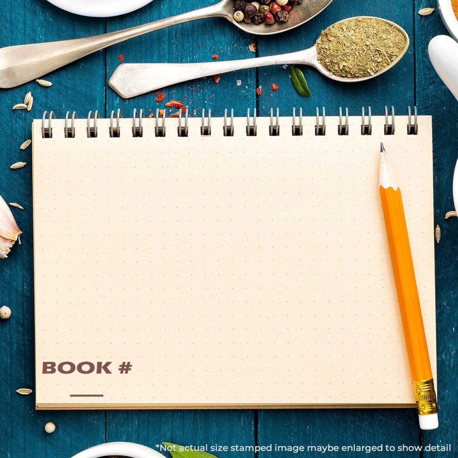 A spiral notebook with a stamped BOOK # using the Book Rubber Stamp, surrounded by spices and a pencil on a blue wooden surface.