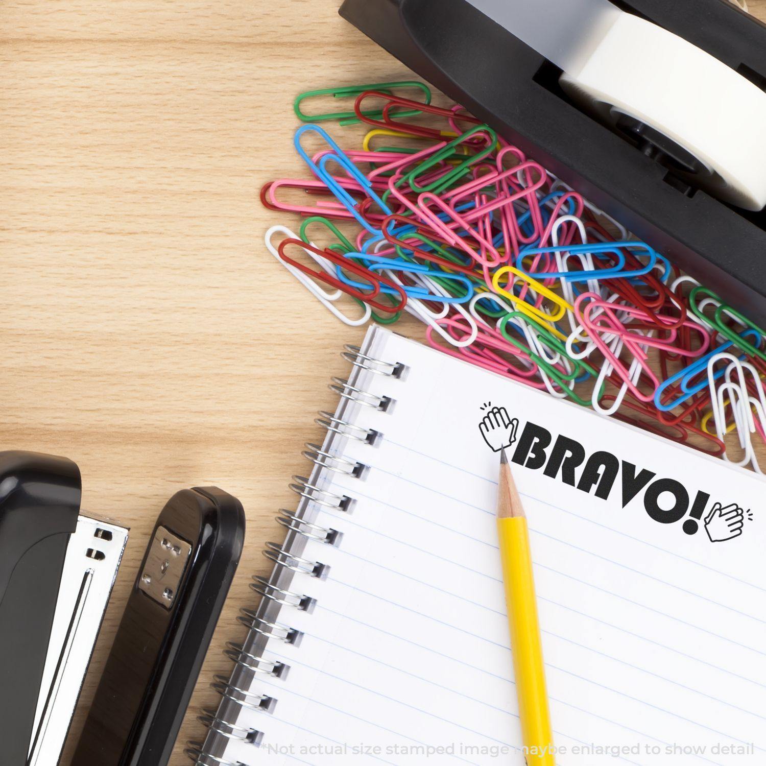 A desk with a stapler, tape dispenser, colorful paperclips, and a notebook stamped with Large Self Inking Bravo with Hands Stamp in bold letters, next to a yellow pencil.