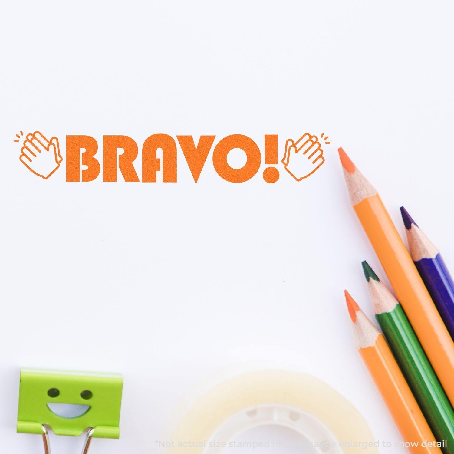 Bravo with Hands Rubber Stamp in orange ink on white paper, surrounded by colored pencils, tape, and a smiling binder clip.