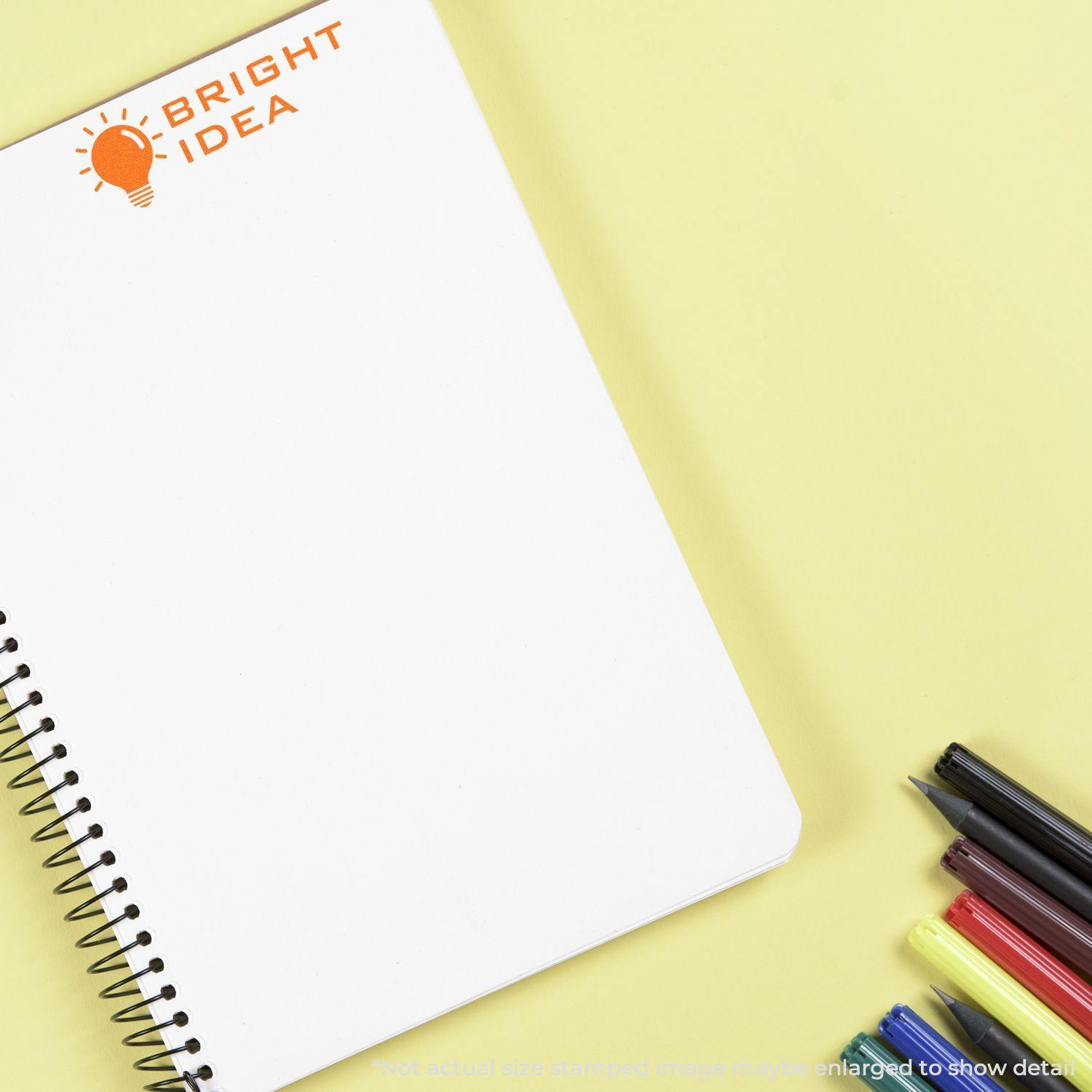 A notebook stamped with the Large Bright Idea Rubber Stamp in orange ink, surrounded by colored pens on a yellow background.