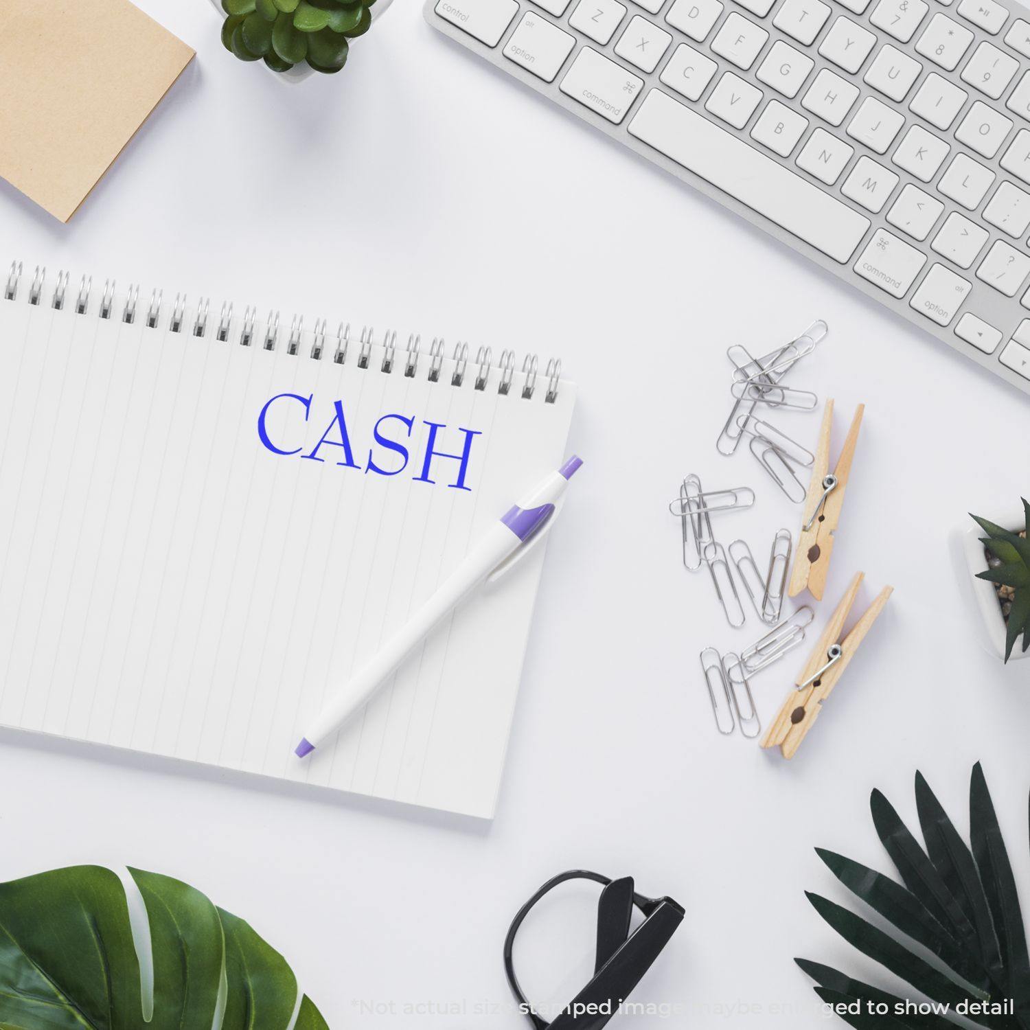 Desk with a notebook stamped with 'CASH' in blue ink, surrounded by a keyboard, pen, plants, and office supplies.