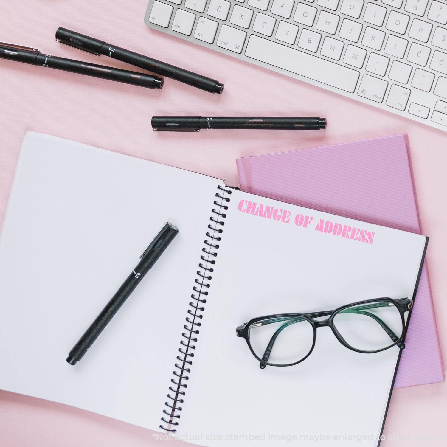 Change Of Address Rubber Stamp used on a notebook, surrounded by pens, glasses, and a keyboard on a pink background.