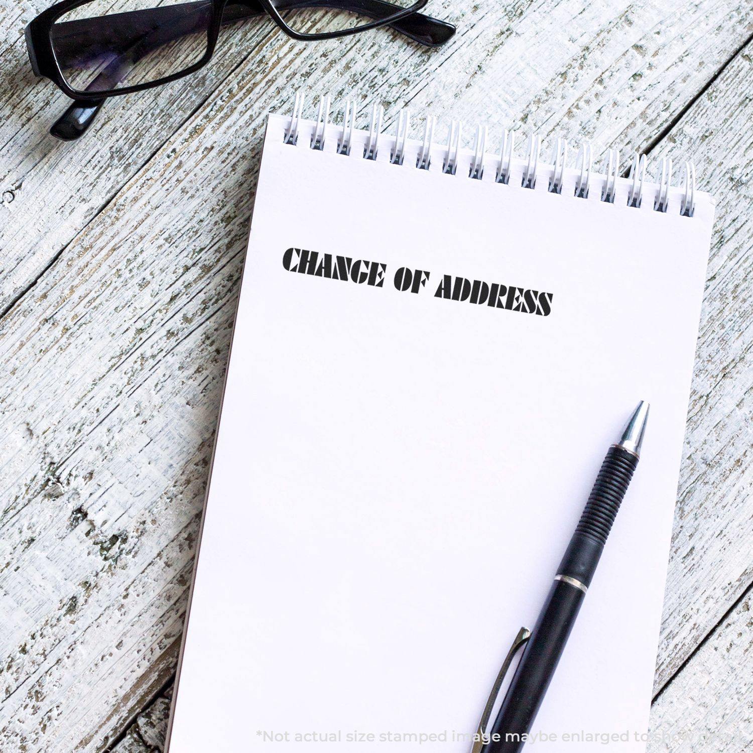 Large Pre-Inked Change Of Address Stamp used on a notepad with CHANGE OF ADDRESS text, next to a pen and glasses on a wooden surface.