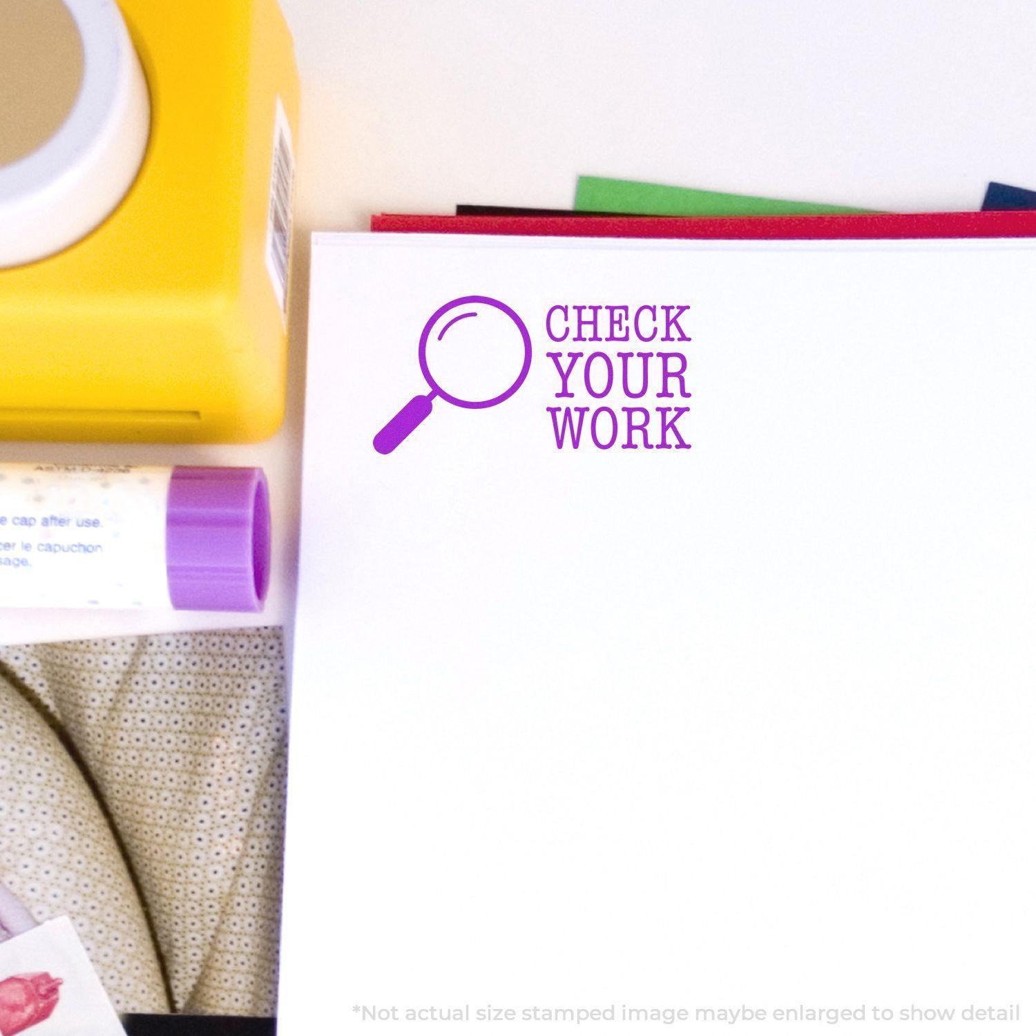 Large Pre-Inked Check Your Work Stamp in purple ink on a white paper, surrounded by office supplies including a yellow container and glue stick.