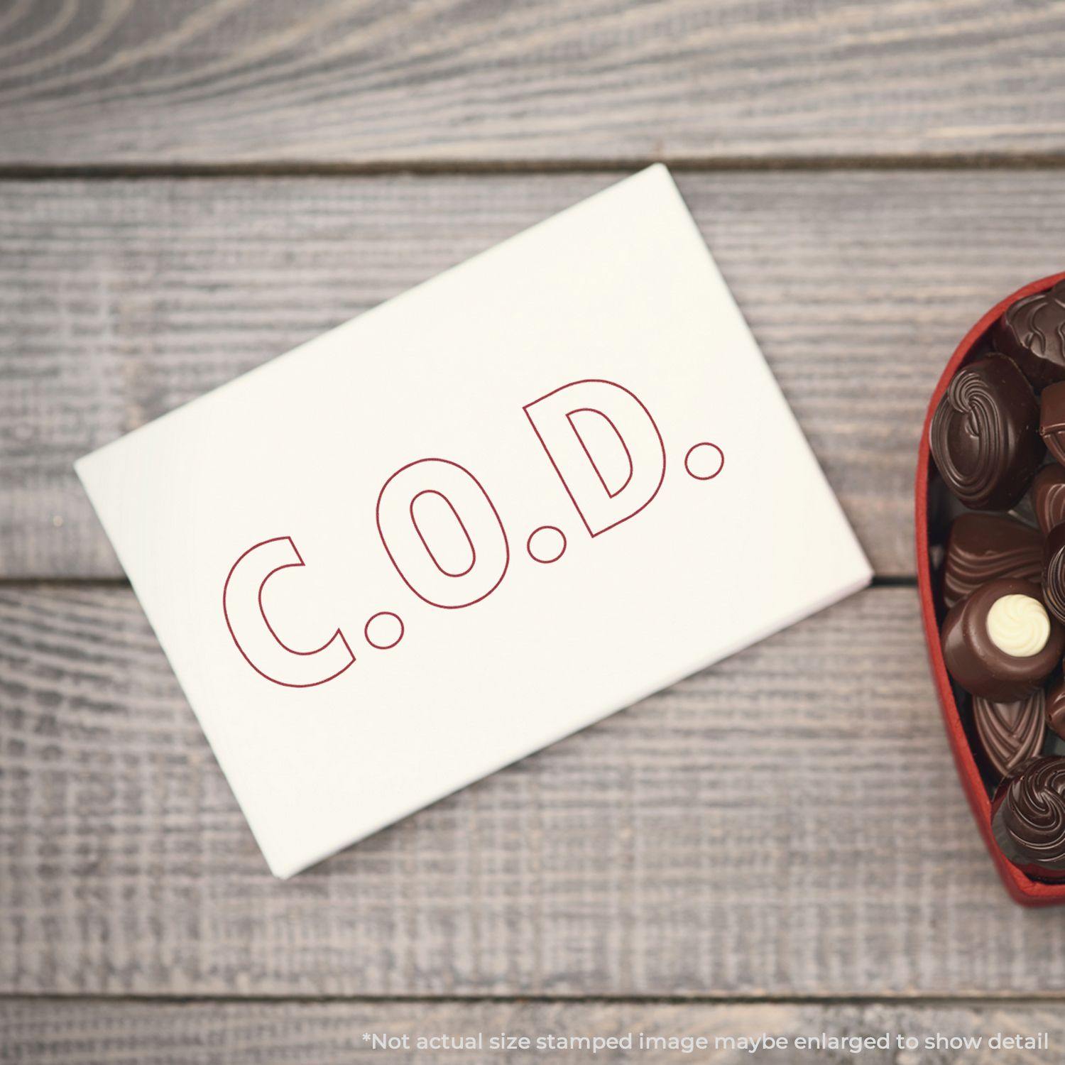 Large Pre-Inked C.O.D. Outline Stamp used on a white card, placed next to a heart-shaped box of chocolates on a wooden surface.