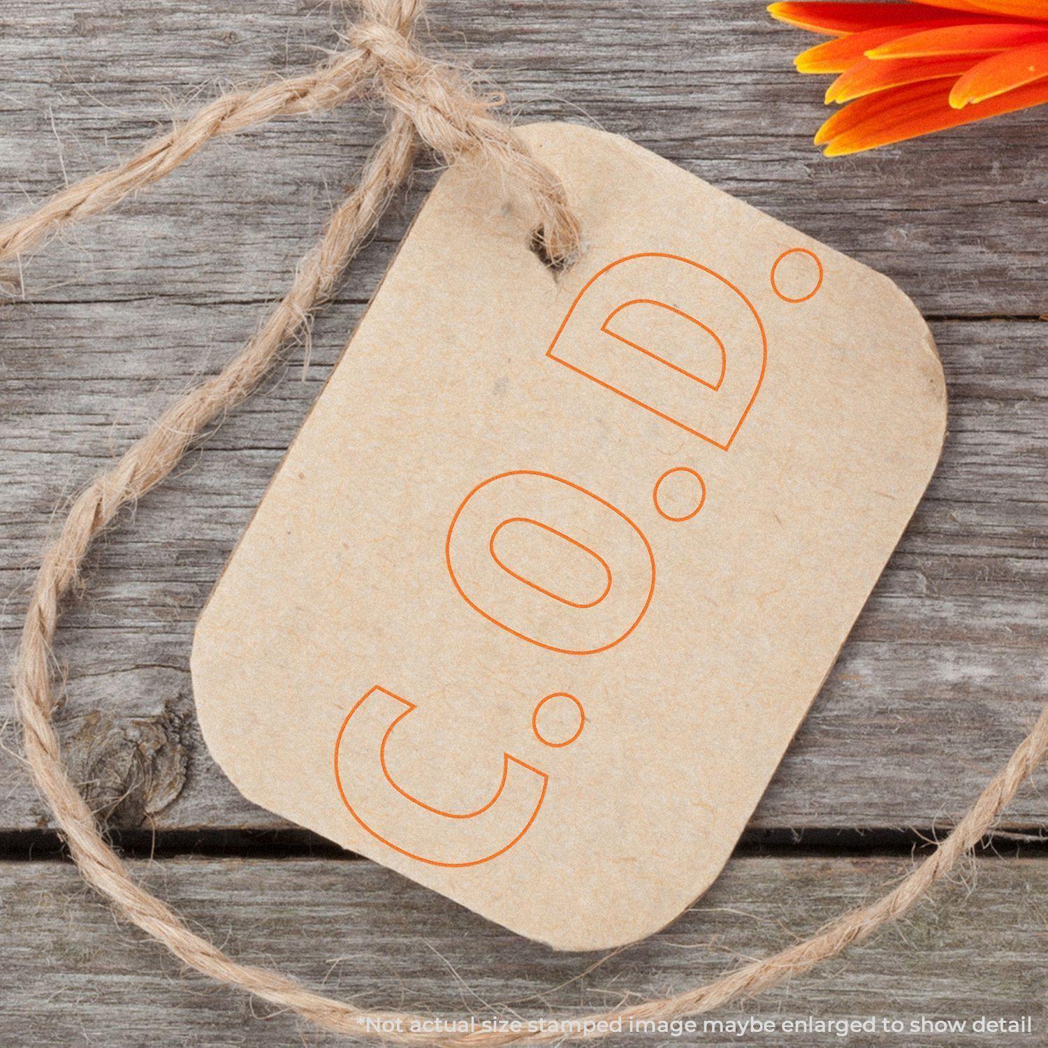 COD With Outline Text Rubber Stamp used on a brown tag with twine, placed on a wooden surface next to an orange flower.