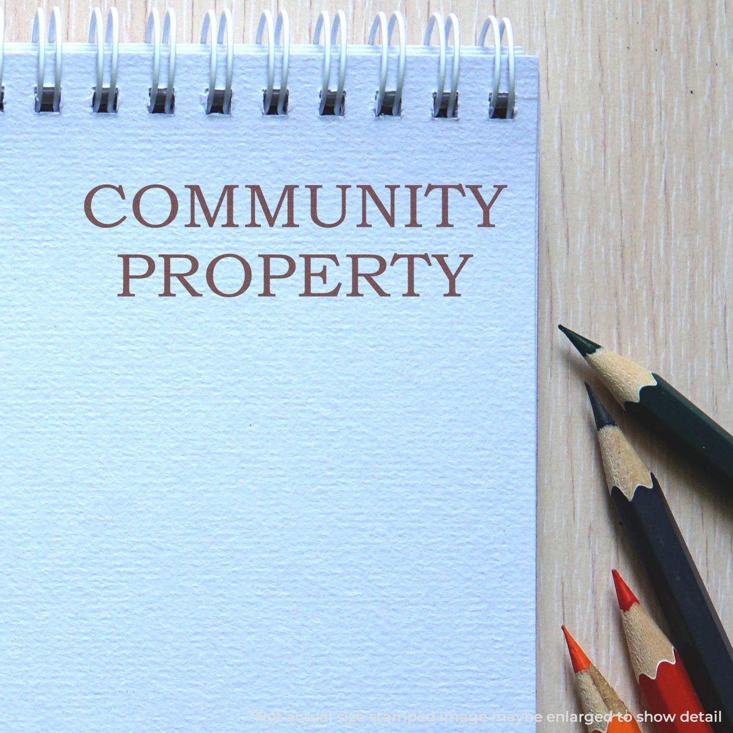 Large Pre-Inked Community Property Stamp used on a spiral notebook with colored pencils nearby on a wooden surface.