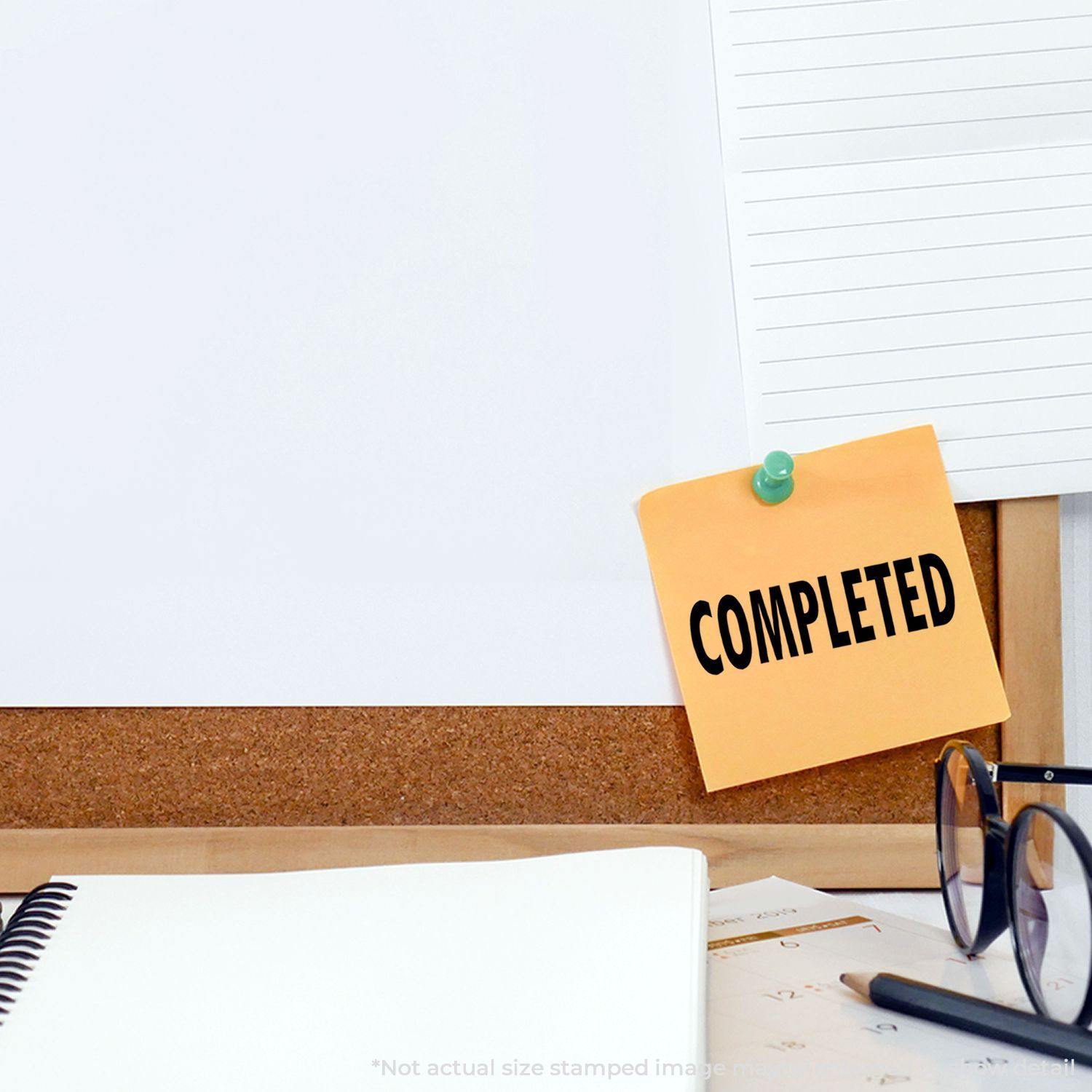 Large Completed Rubber Stamp used on a yellow sticky note pinned to a corkboard, next to a notebook, glasses, and a calendar.