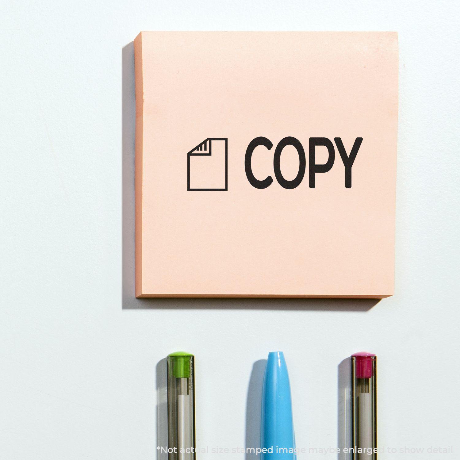 A pink sticky note stamped with COPY using the Copy with Letter Rubber Stamp, placed above three pens on a white surface.