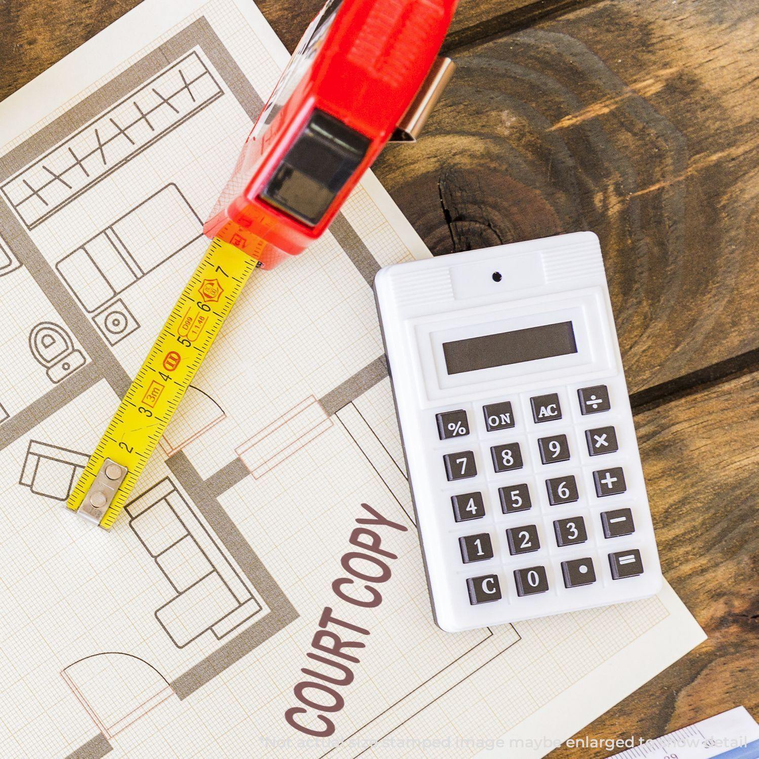 Large Court Copy Rubber Stamp next to a calculator, tape measure, and architectural plan on a wooden surface.