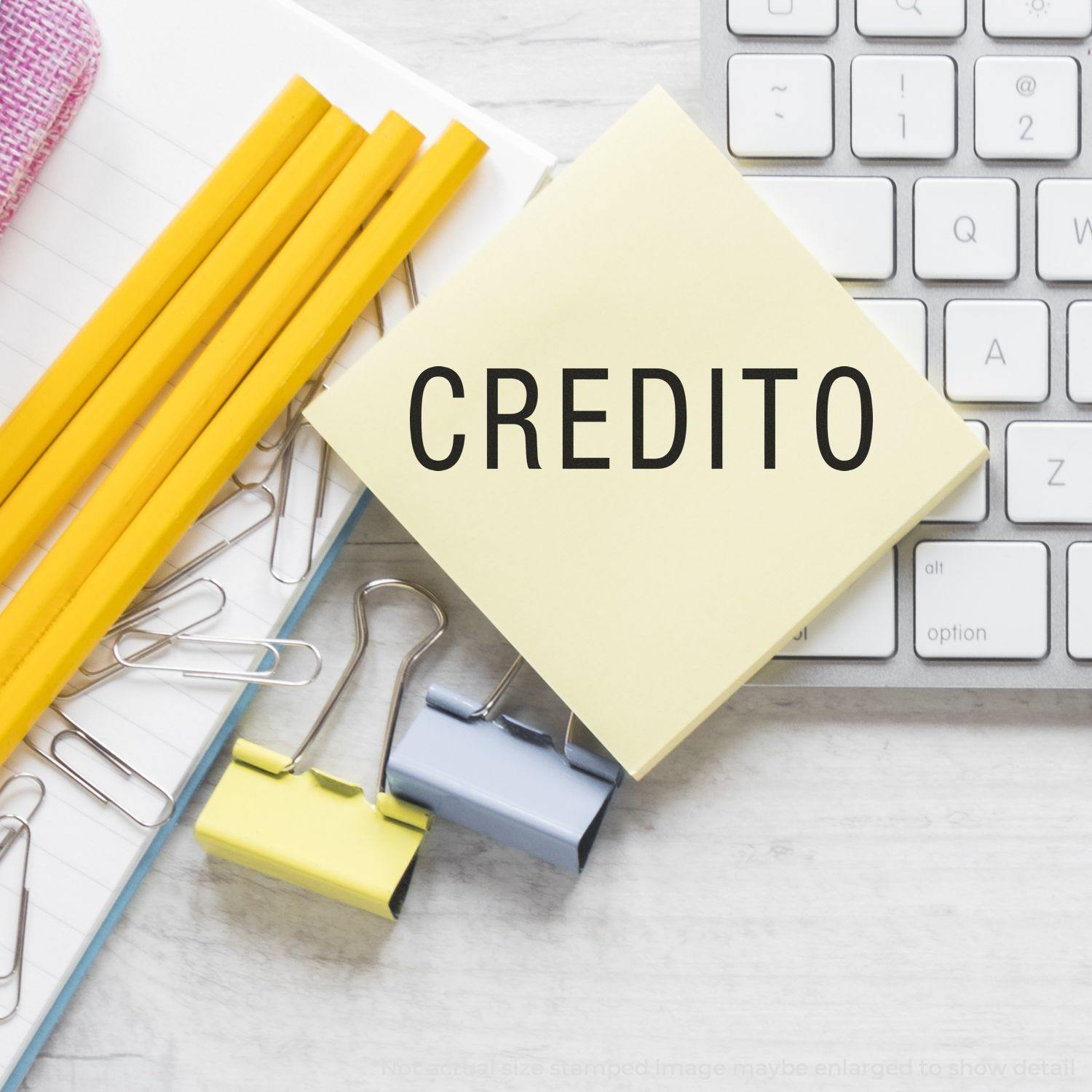 Self Inking Credito Stamp on a yellow sticky note next to pencils, paper clips, binder clips, and a keyboard on a desk.