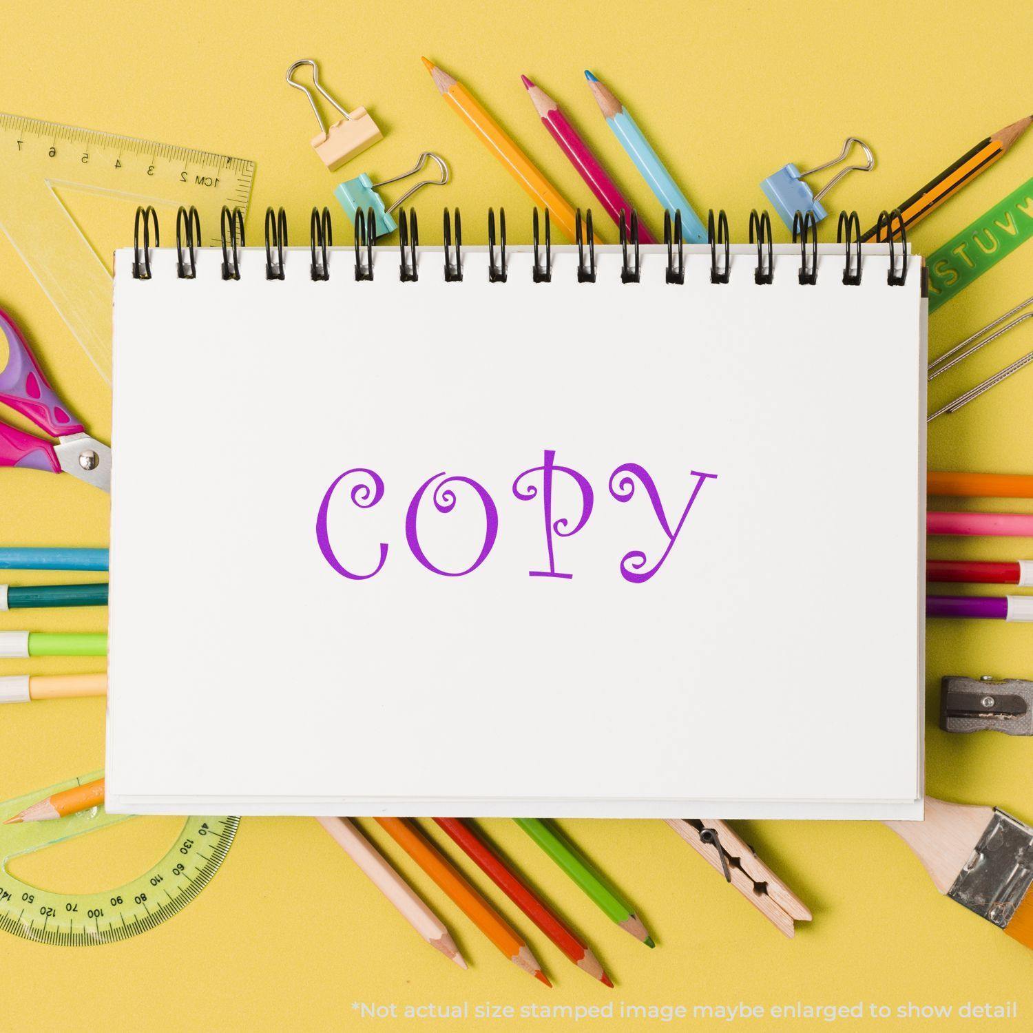 Large Pre-Inked Curly Copy Stamp imprint on a white notebook surrounded by colorful pencils, rulers, and other stationery on a yellow background.