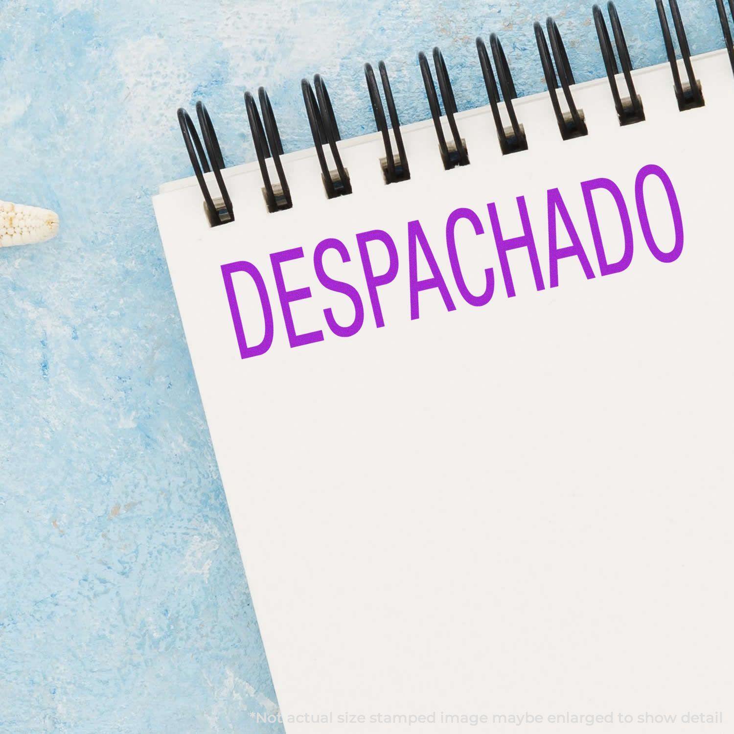 A notebook with DESPACHADO stamped in purple ink using the Despachado Rubber Stamp, placed on a light blue textured surface.