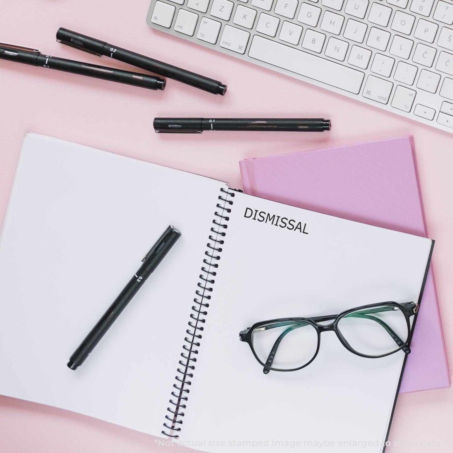 Self Inking Dismissal Stamp used on a notebook, surrounded by pens, glasses, a keyboard, and a pink folder on a pink desk.