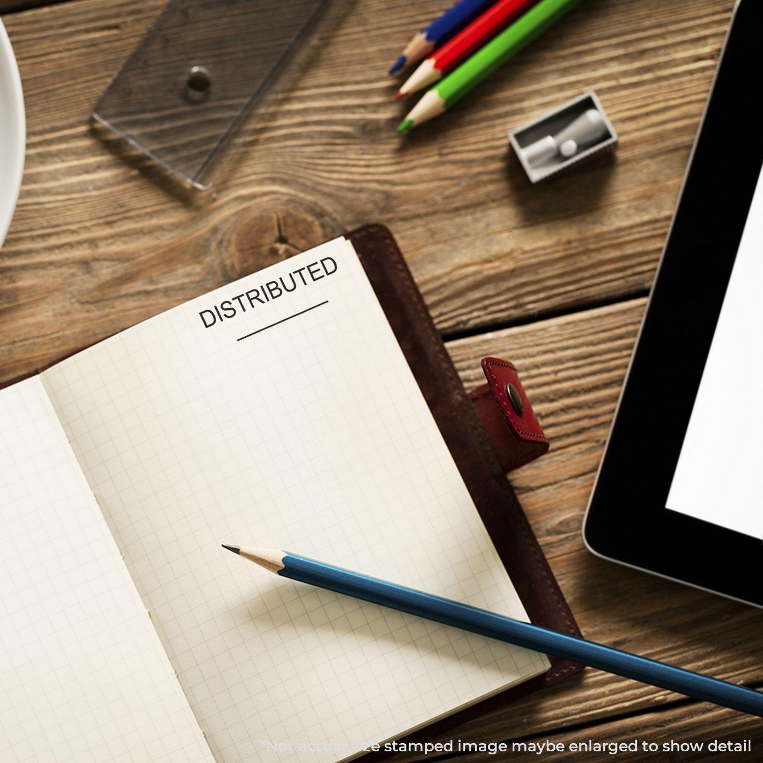 Distributed Rubber Stamp used on a notebook page, surrounded by pencils, a sharpener, and a tablet on a wooden desk.